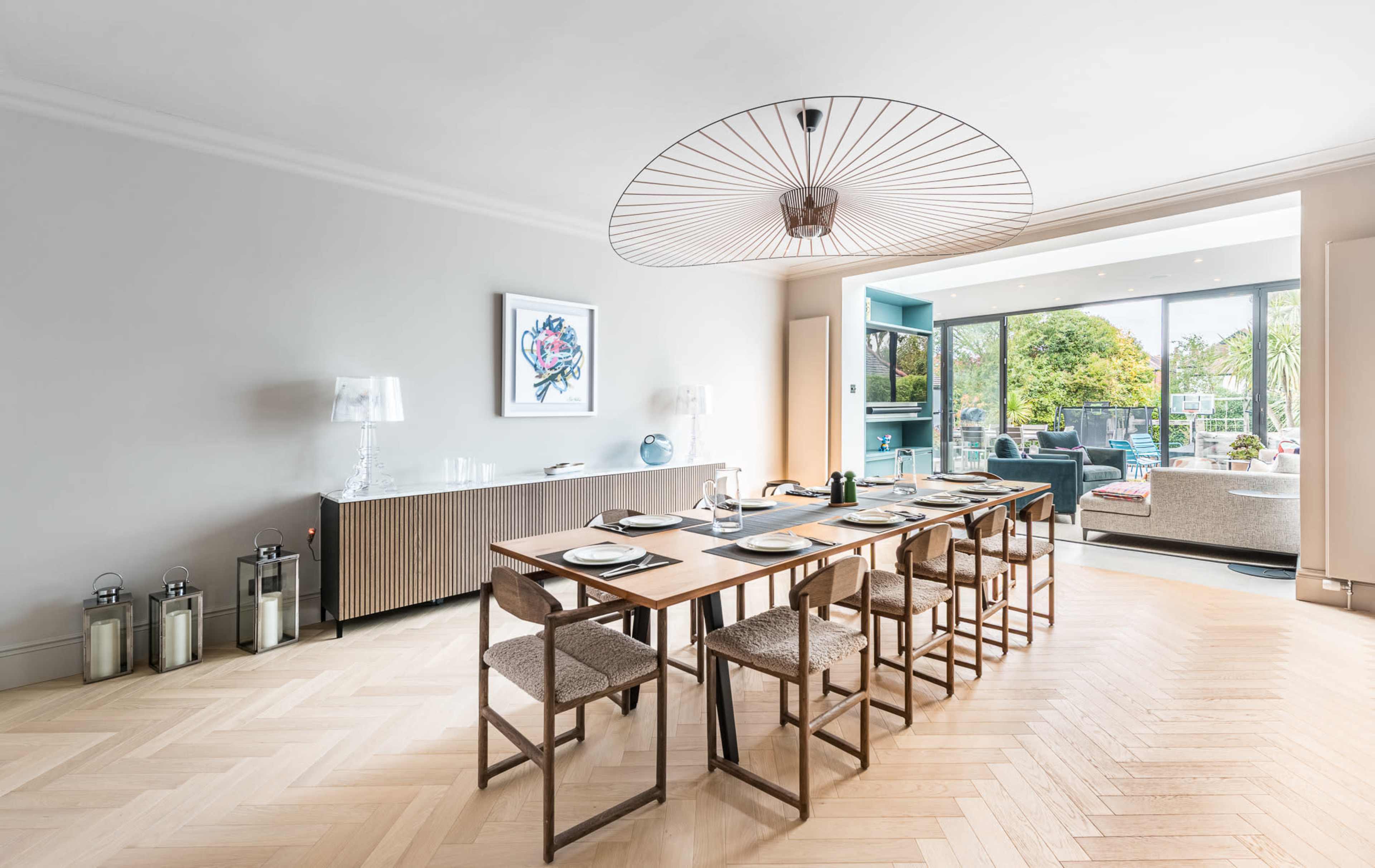 A modern dining area features a long wood table surrounded by chairs, with a large light fixture above and a view of a garden through glass doors.