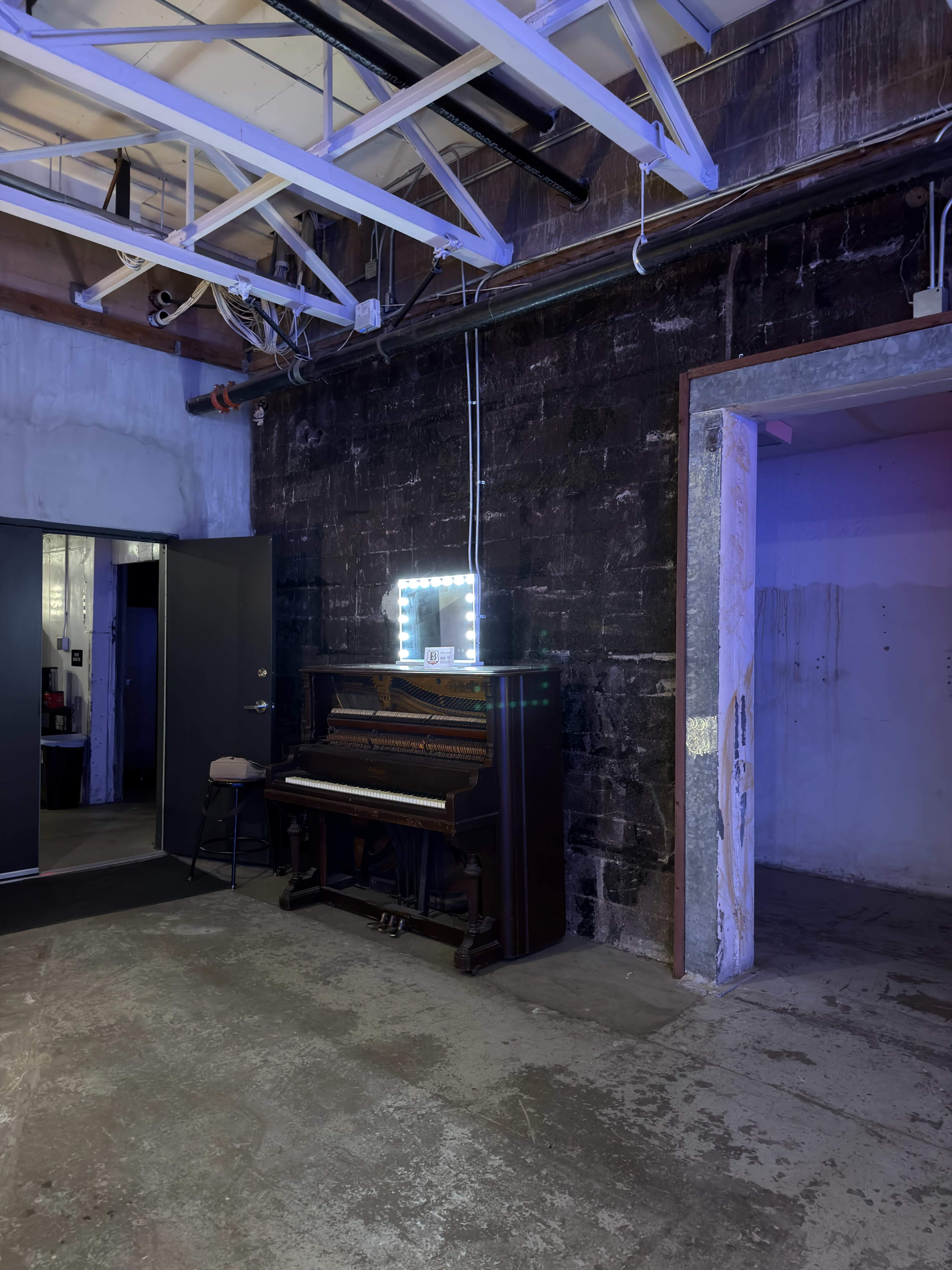 A vintage piano is positioned against a stark black wall, next to a brightly lit mirror, in an industrial-style room with concrete flooring.