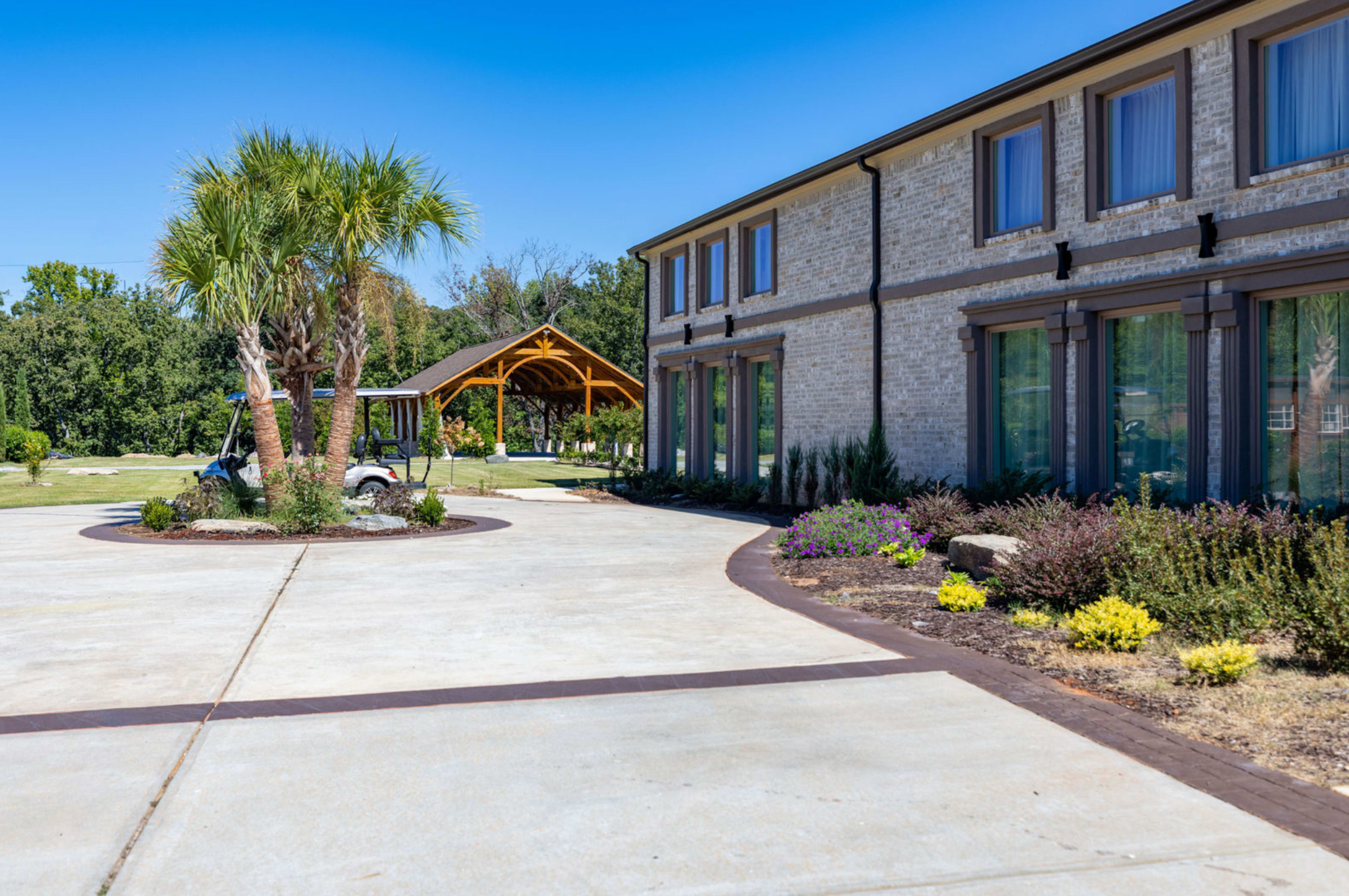 The image shows a landscaped property with a building on one side, a curved driveway, and a wooden gazebo in the background.