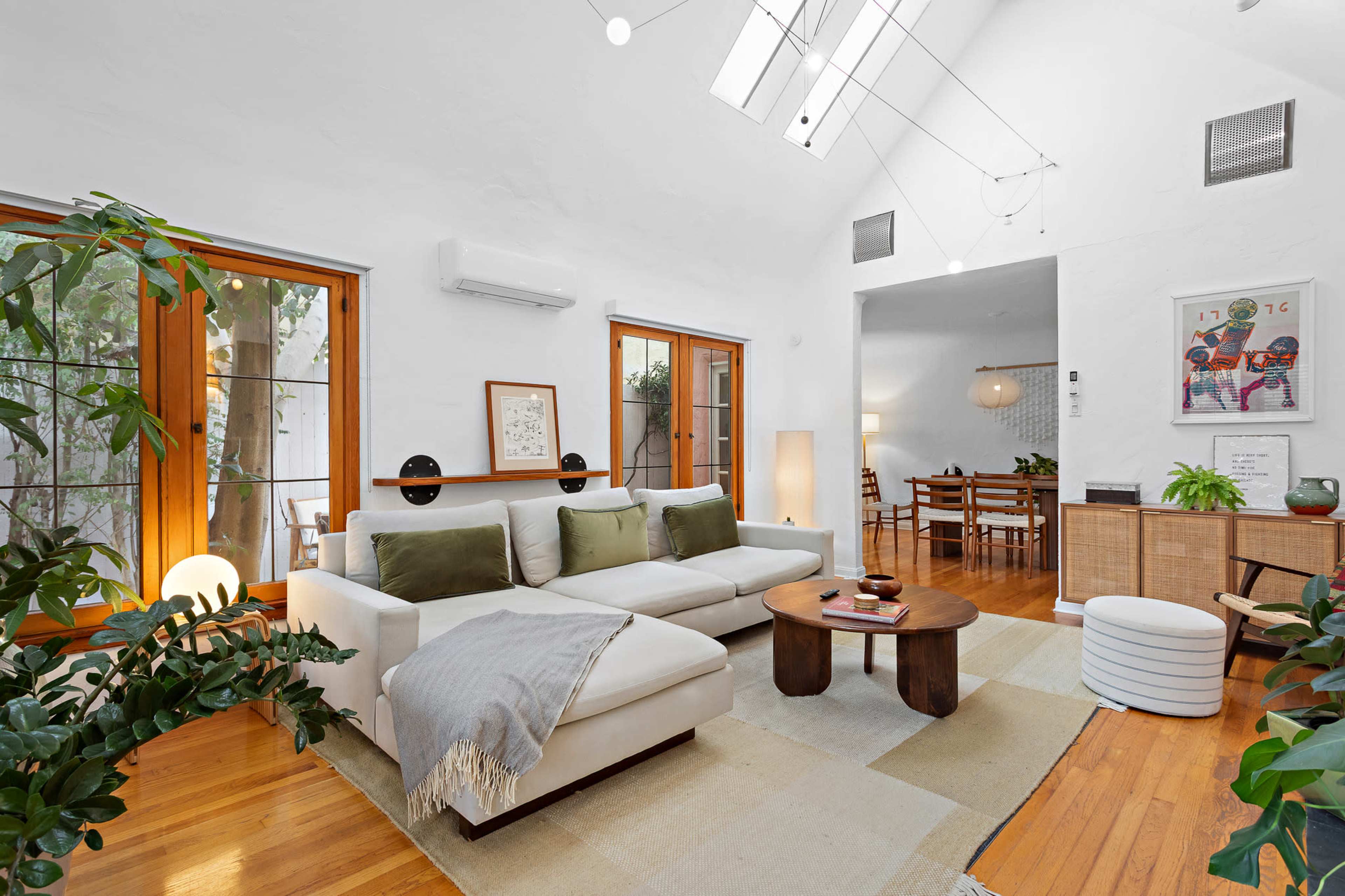 The image shows a bright living room featuring a white sectional sofa, a round coffee table, and large windows that allow natural light to fill the space.