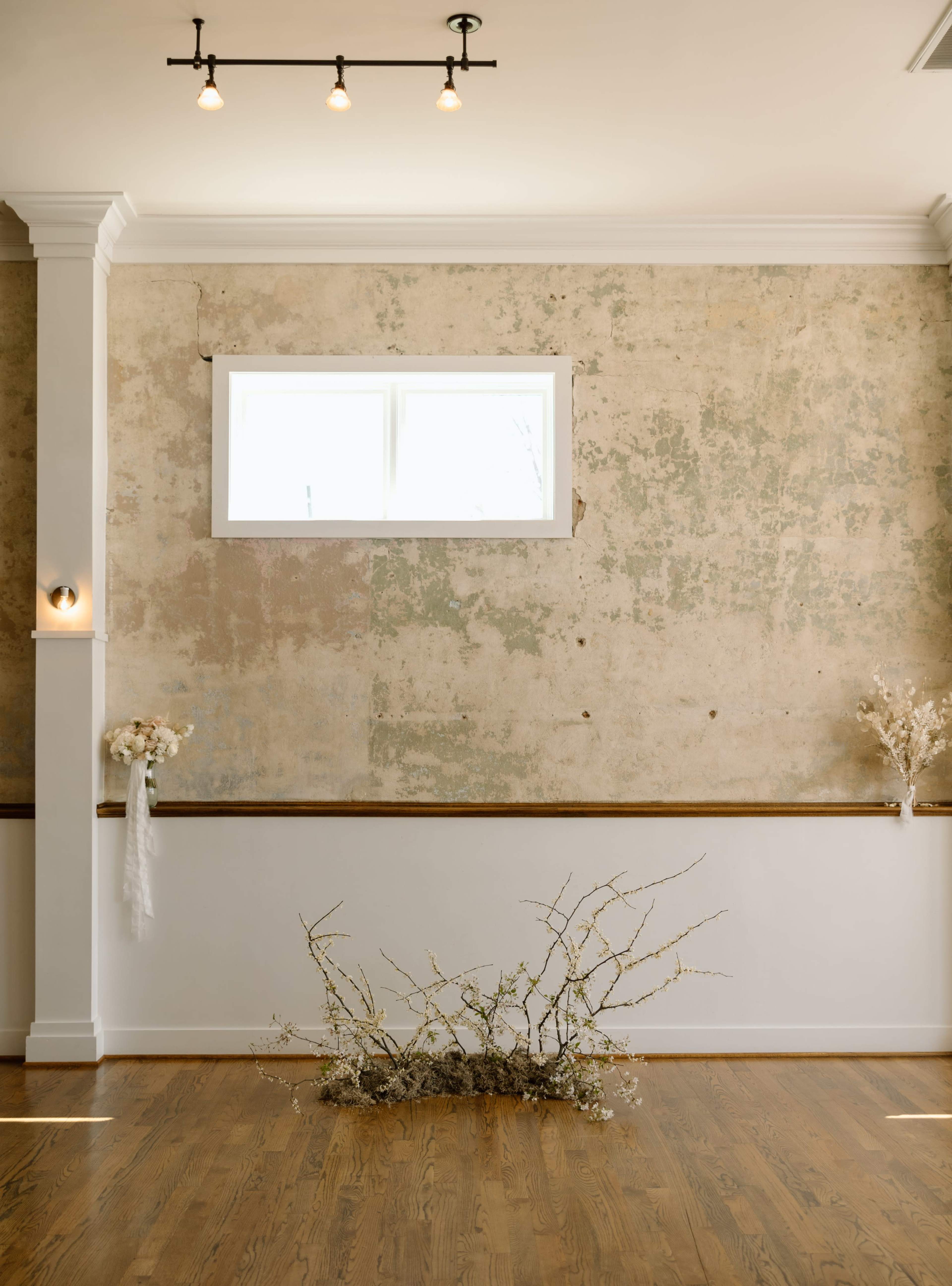 A simple interior features a weathered wall, a window, and natural elements arranged on a wooden floor.