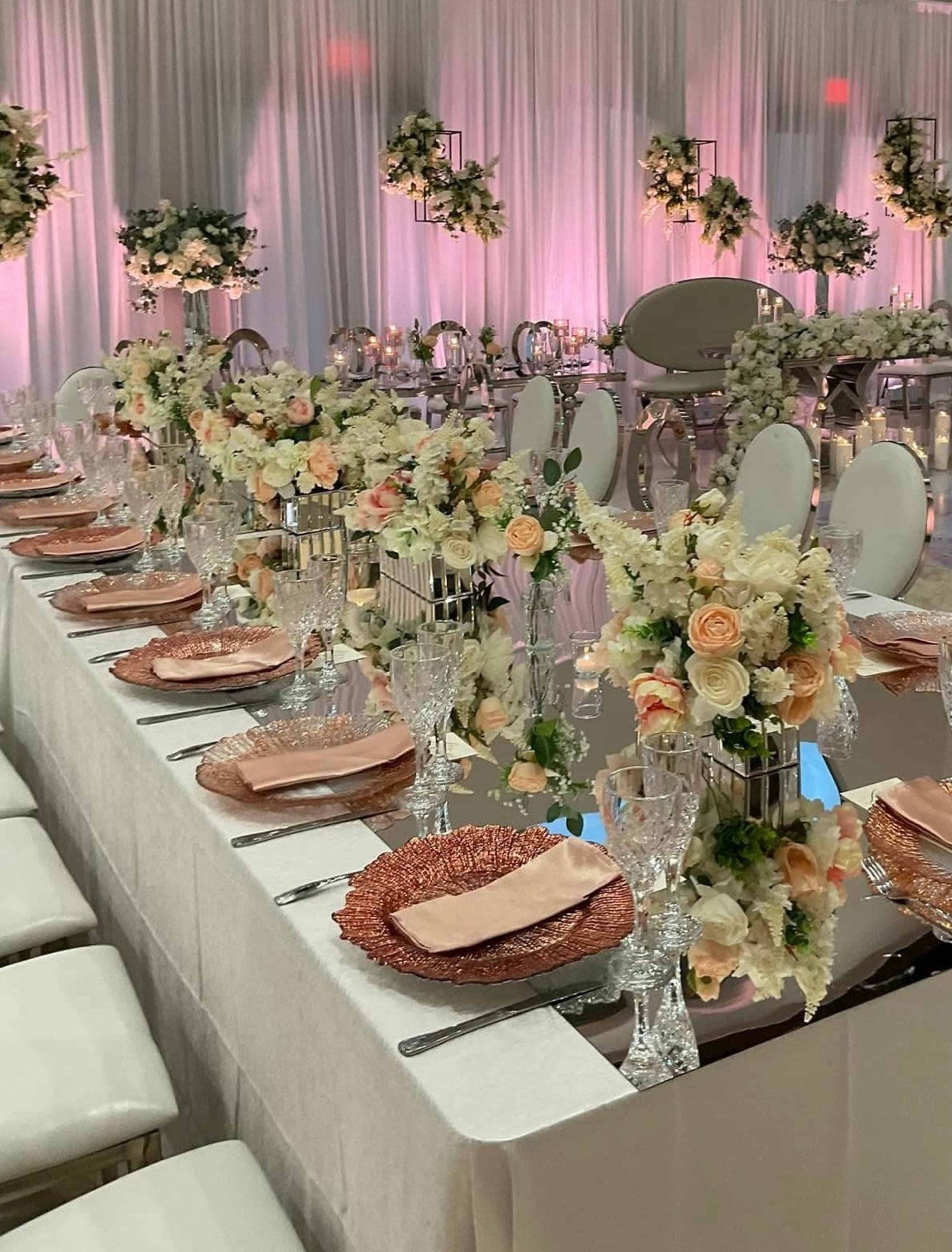A elegantly arranged banquet table is set with delicate floral centerpieces, glassware, and fine china, all under soft lighting and draped fabric.