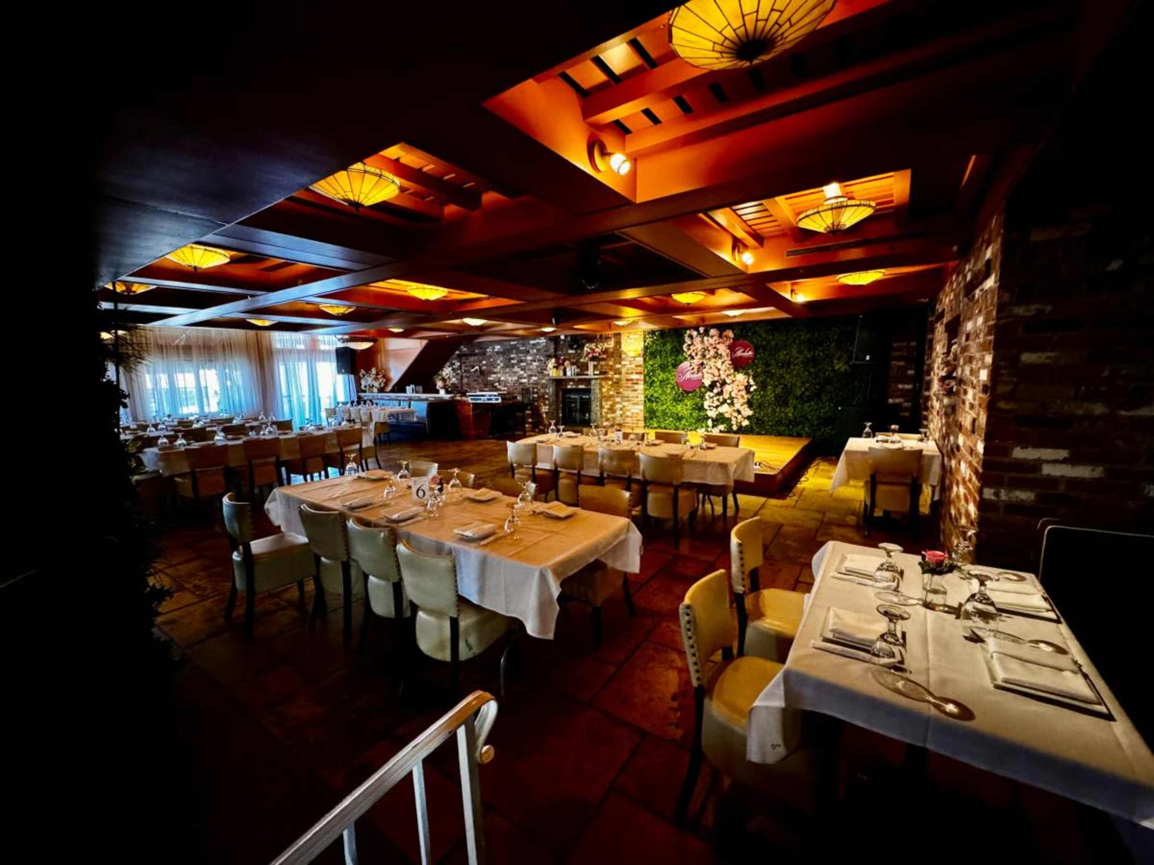The image shows an elegantly arranged restaurant interior featuring tables with white tablecloths, ornate lighting, and a decorative wall with plants.