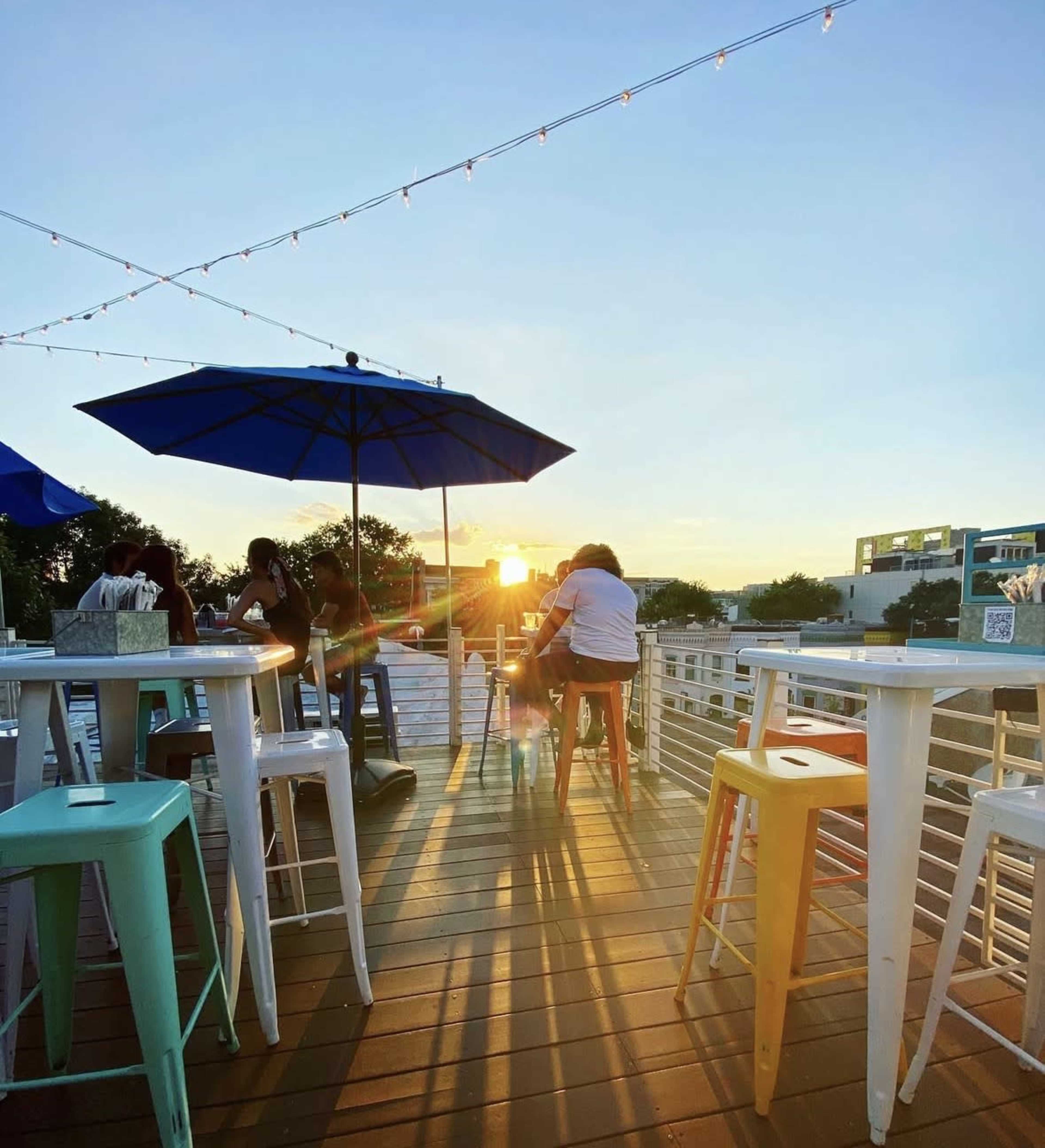 The image shows a rooftop bar setting at sunset, with patrons sitting at white and colorful stools under umbrellas.