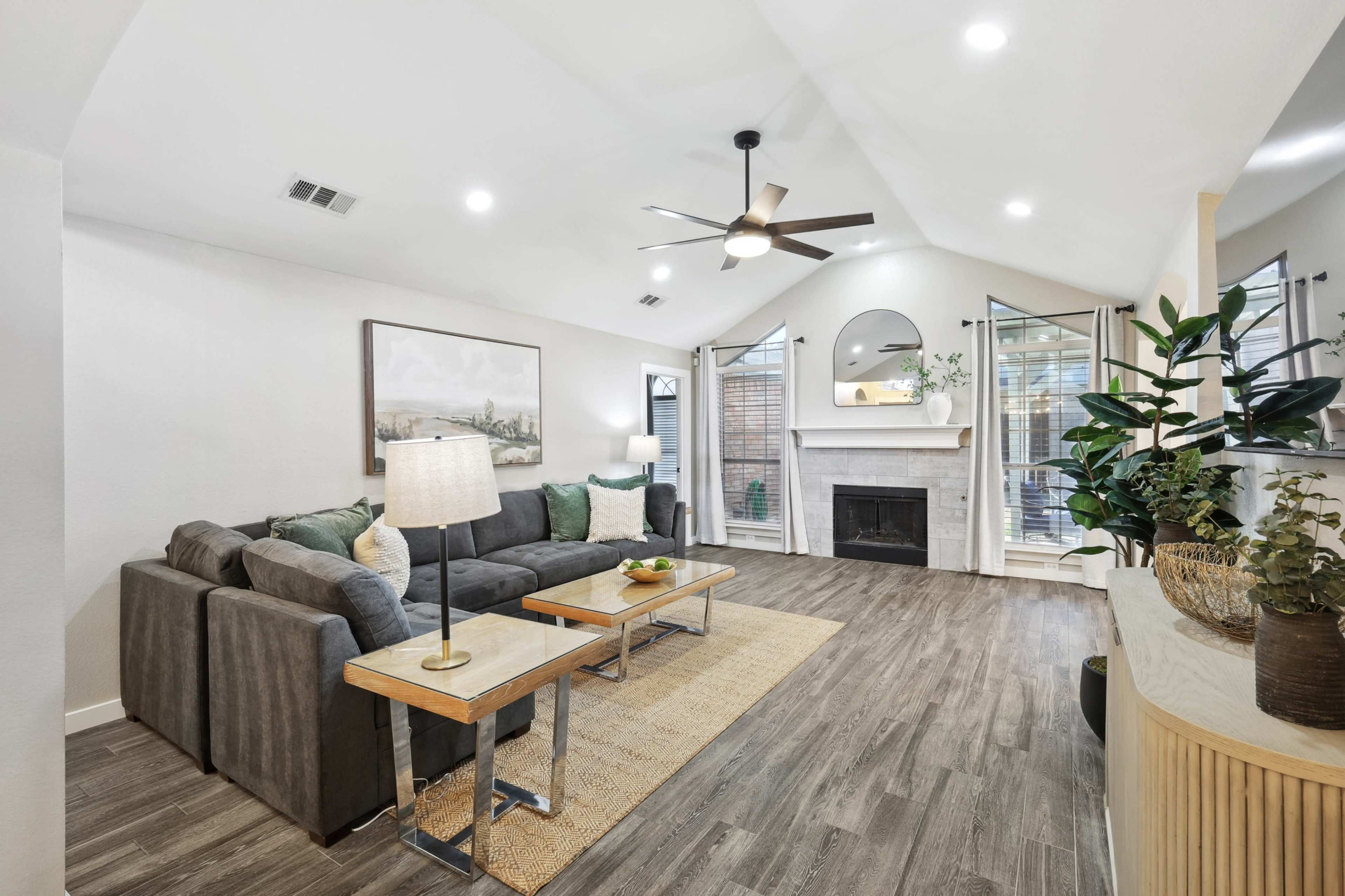 A modern living room features a gray sectional sofa, wooden side tables, a fireplace, and large windows allowing natural light.