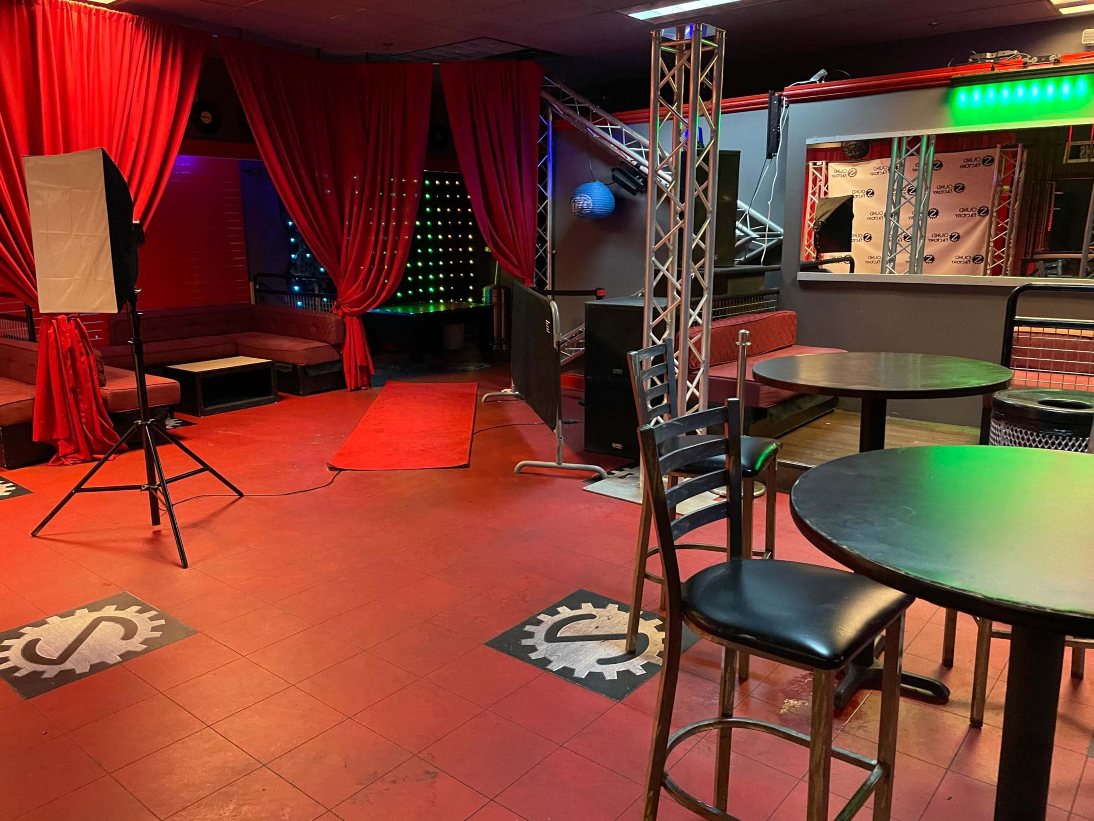 An empty nightclub interior with red curtains, a small stage area, and tables with chairs in a dimly lit setting.