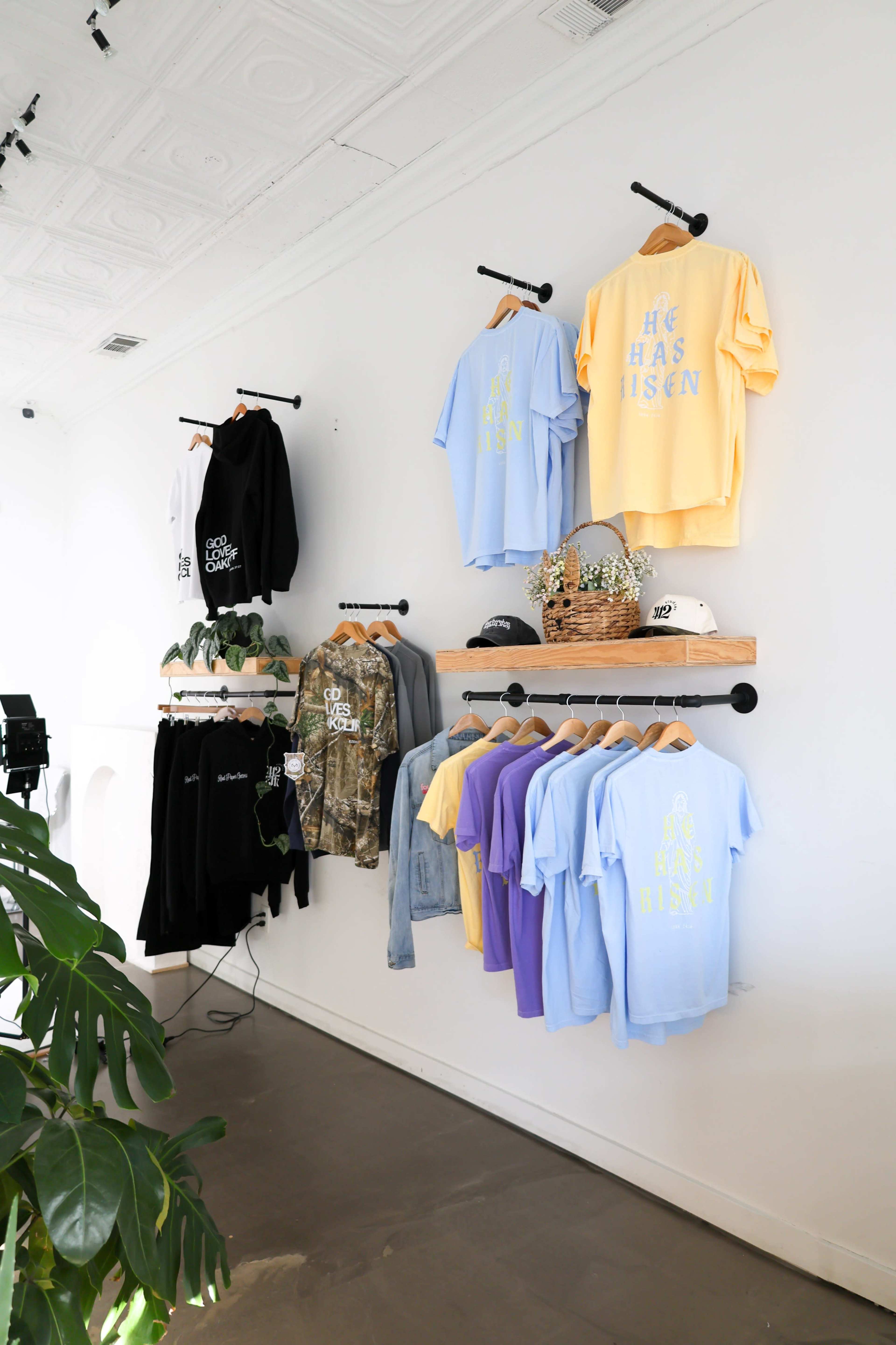 The image shows a clothing display on a white wall featuring various t-shirts in pastel colors, a camouflage hoodie, and a baseball cap, arranged on wooden shelves and hanging rods.