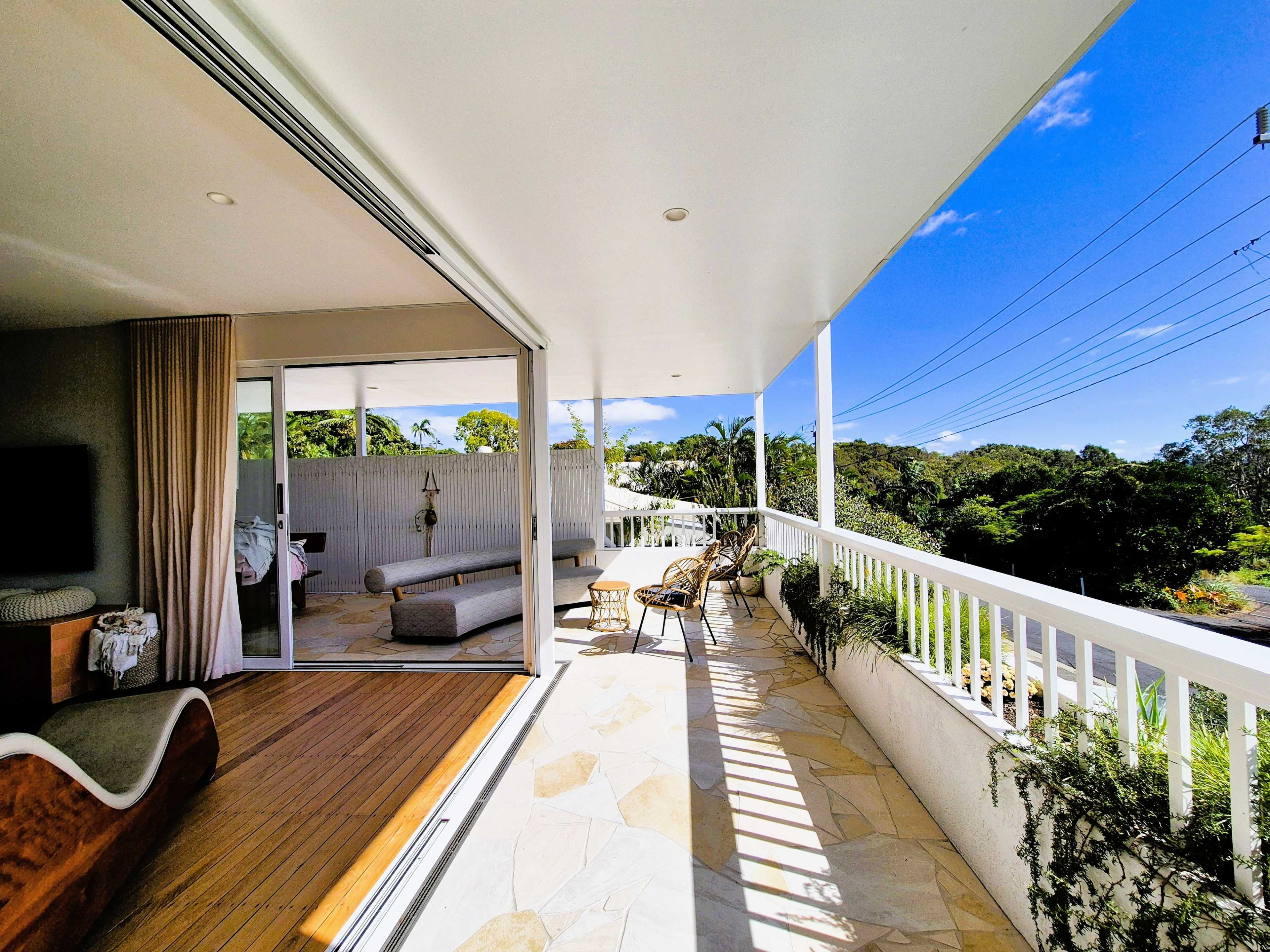 The image shows a spacious terrace with outdoor seating, extending from a modern interior area, surrounded by greenery and under a clear blue sky.