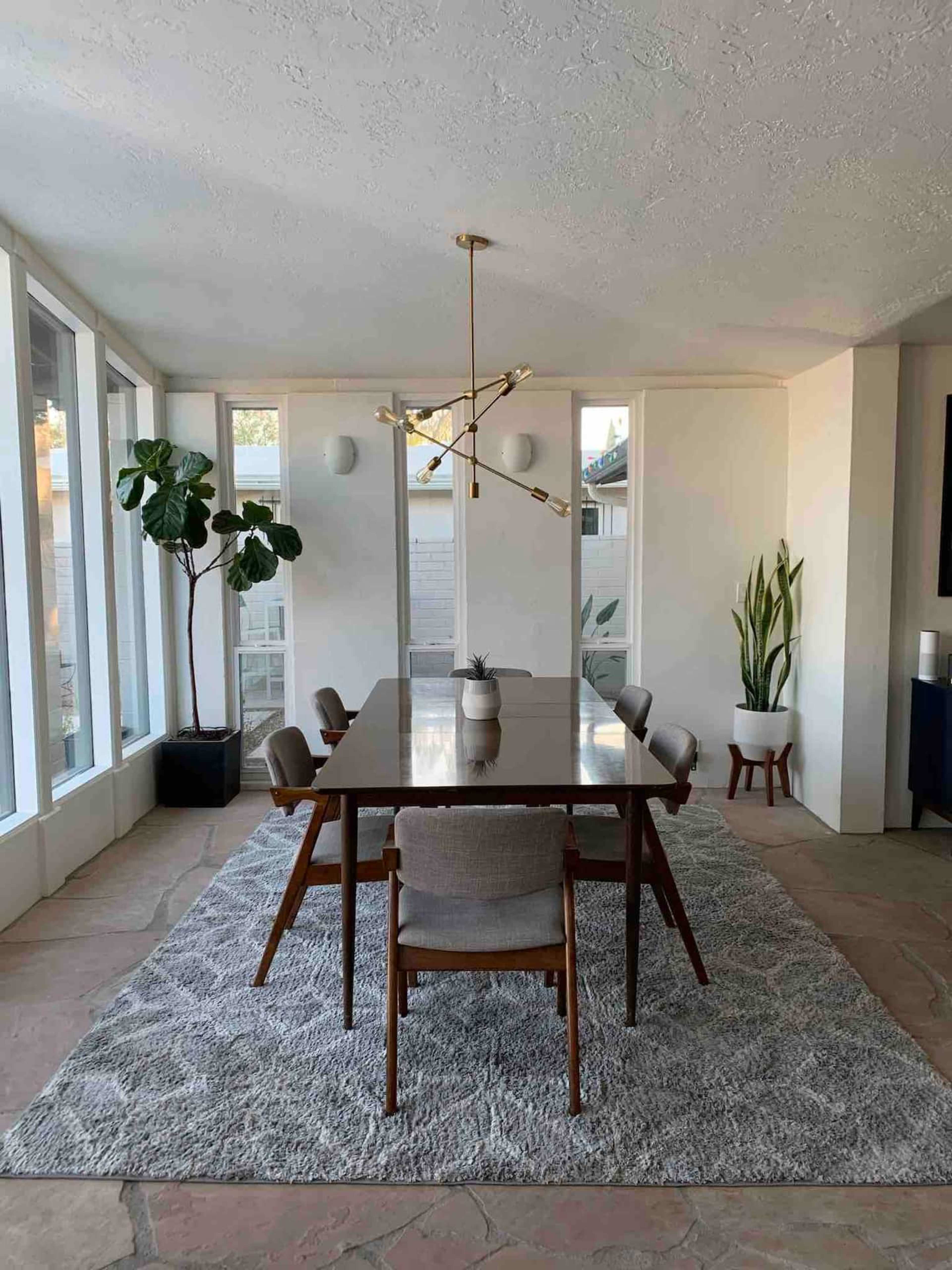A dining room features a long table surrounded by chairs, with large windows allowing natural light, plants in the corners, and a modern chandelier overhead.