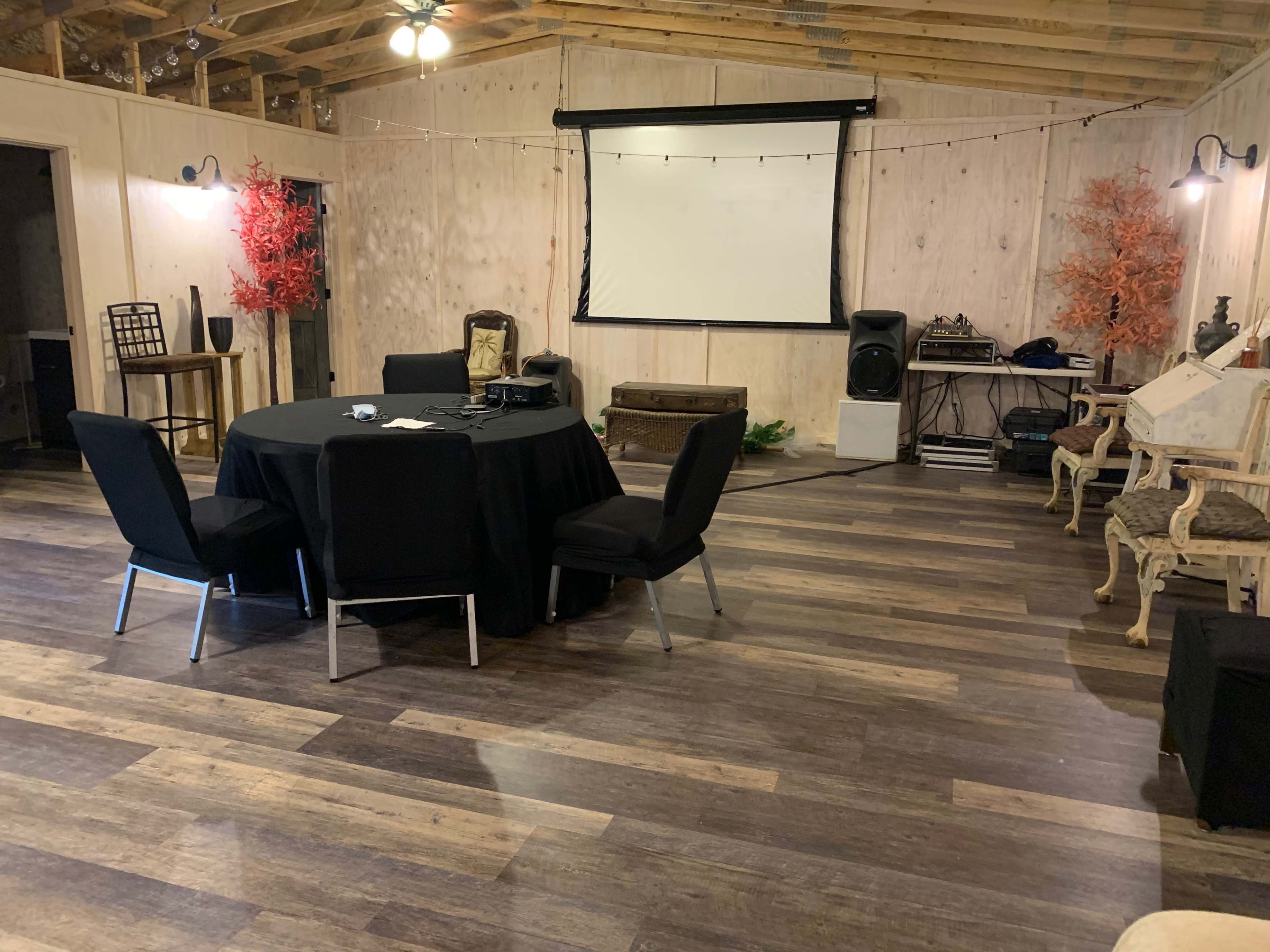 The interior of a room features a round table with dark chairs, a projector screen, and wooden flooring, accompanied by decorative elements and audio equipment.