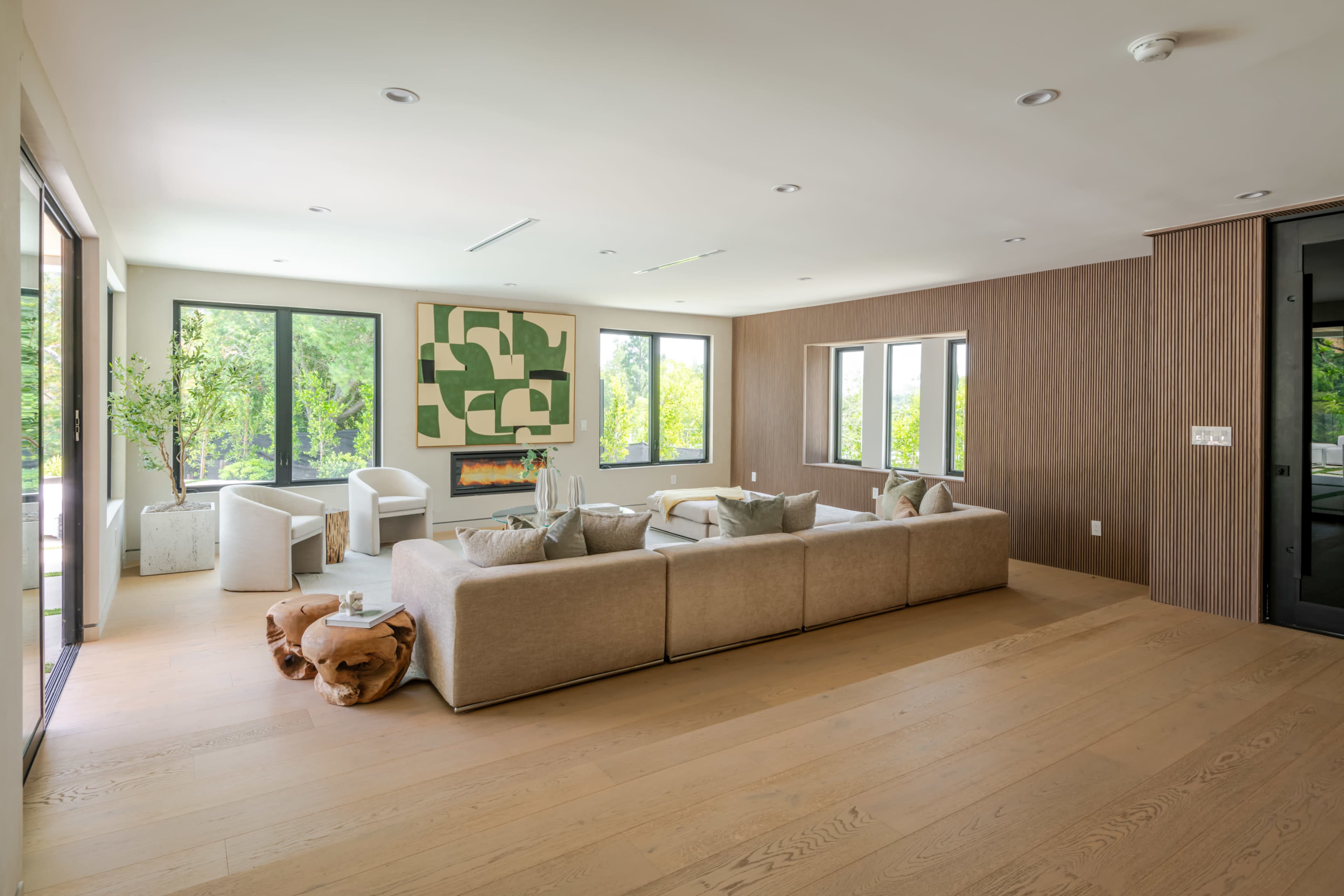 The living room features a sectional sofa, large windows providing natural light, and wooden accents on the walls and floor.