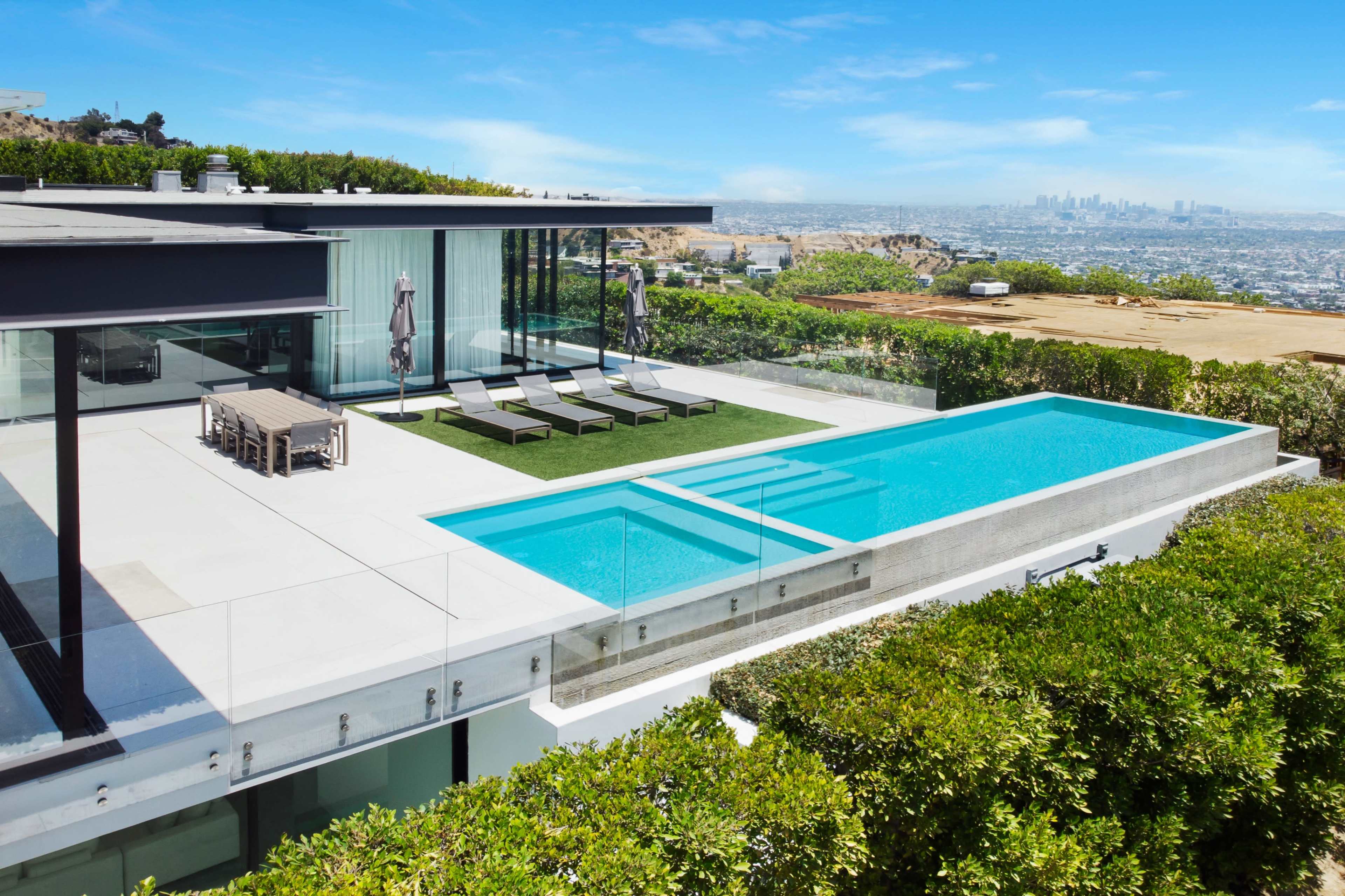 A modern house features a sleek swimming pool and a landscaped terrace with lounge chairs, overlooking a city skyline.