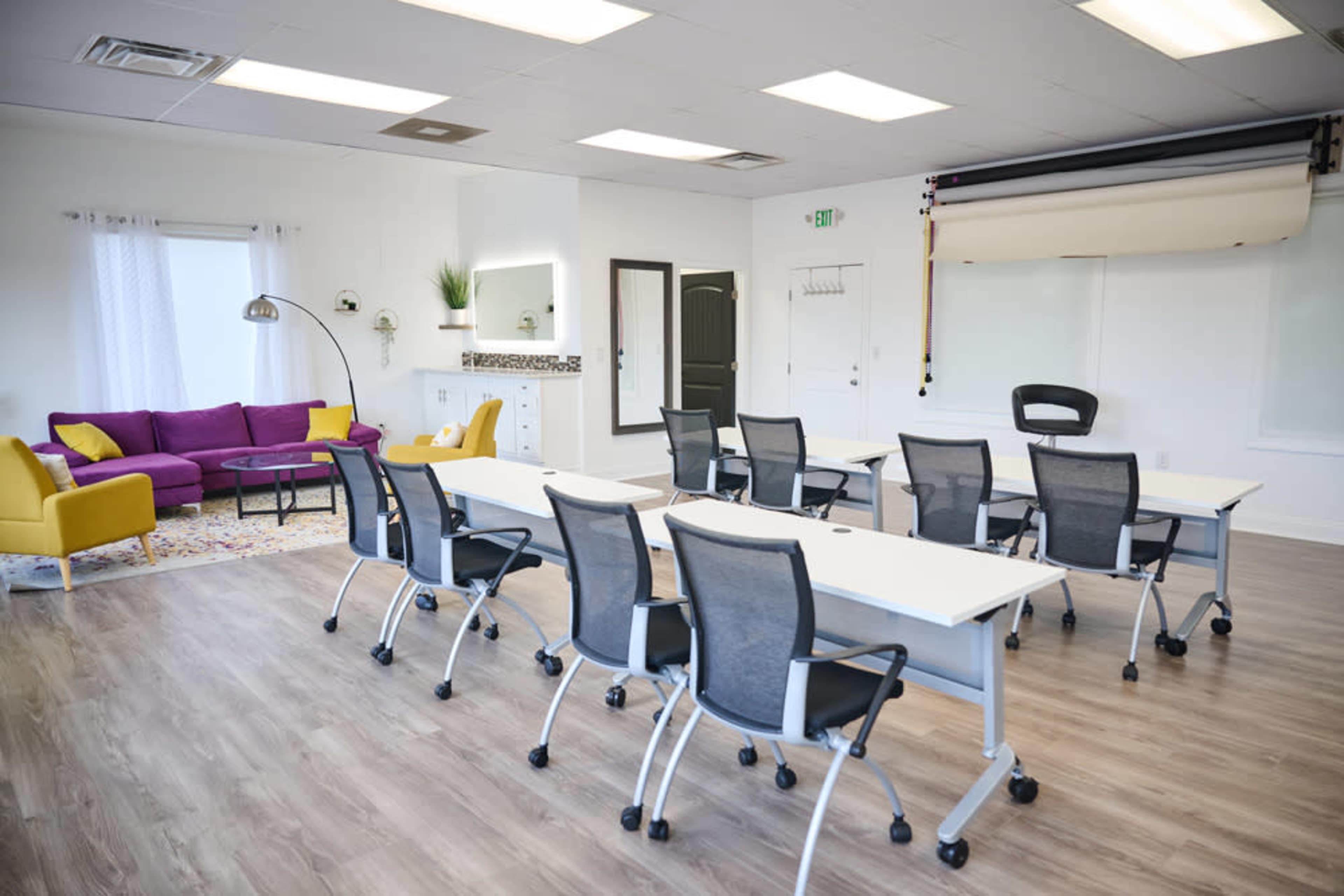 The image shows a bright, modern office space featuring several tables and chairs arranged for a meeting, with a seating area that includes a purple couch and a yellow chair.