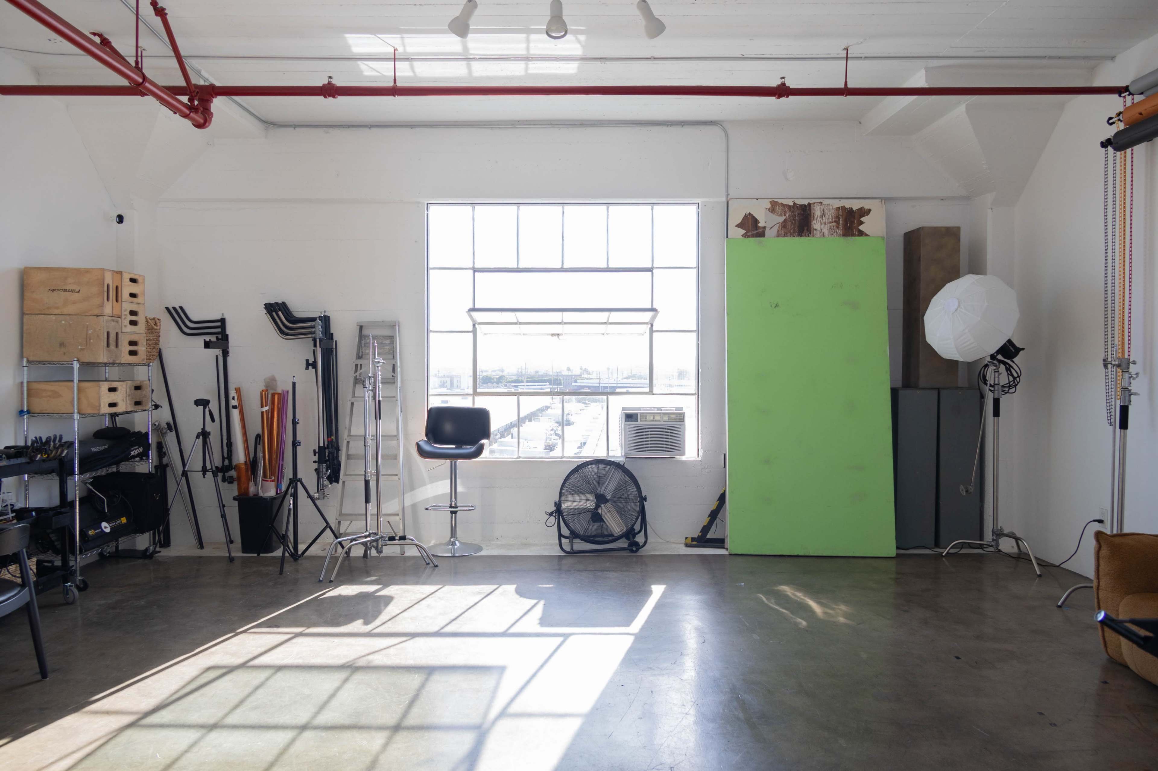 The image shows a spacious studio featuring a large window, a green backdrop, photography equipment, and a bar stool.