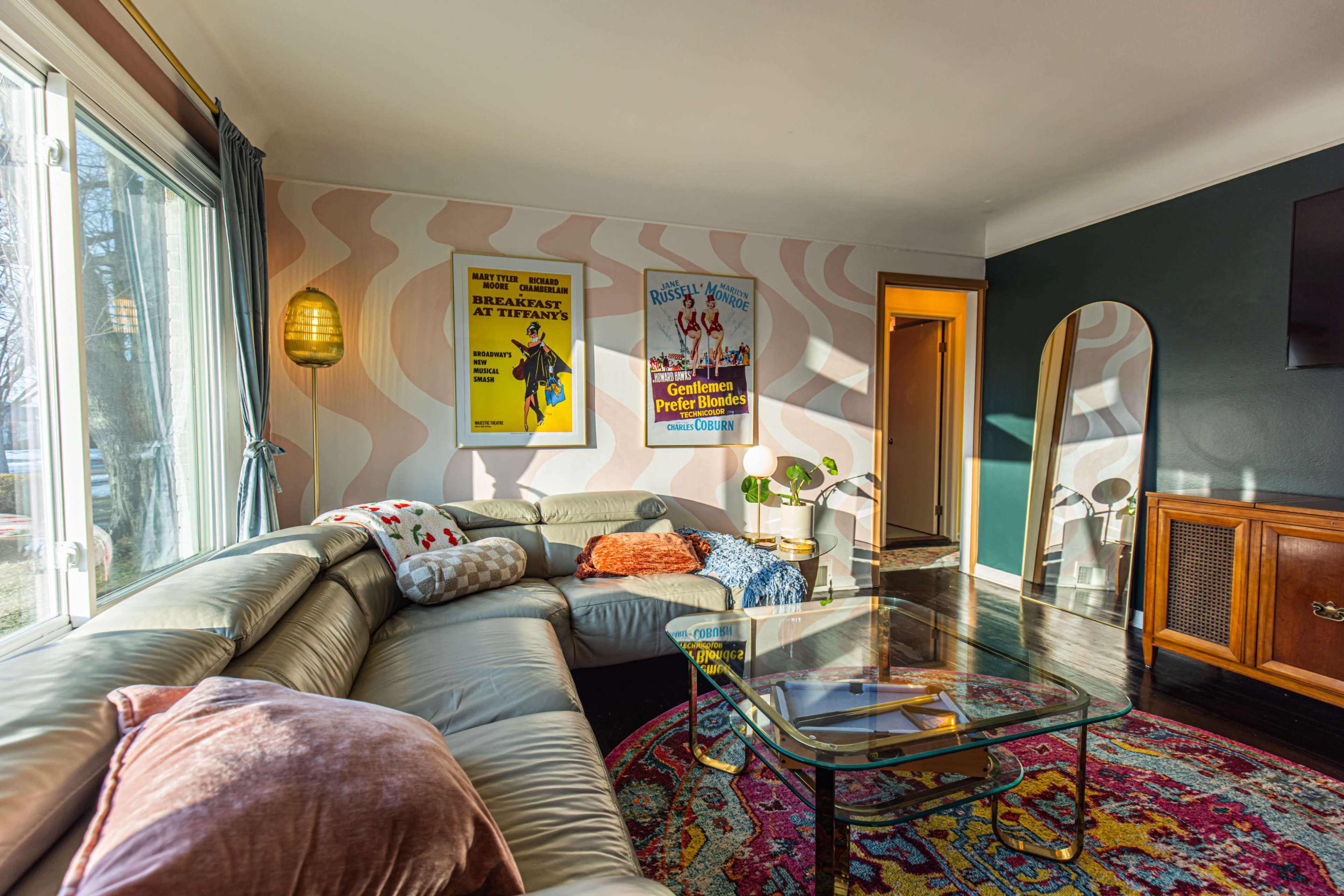 A cozy living room features a sectional sofa, a glass coffee table, retro movie posters on the walls, and large windows that let in natural light.