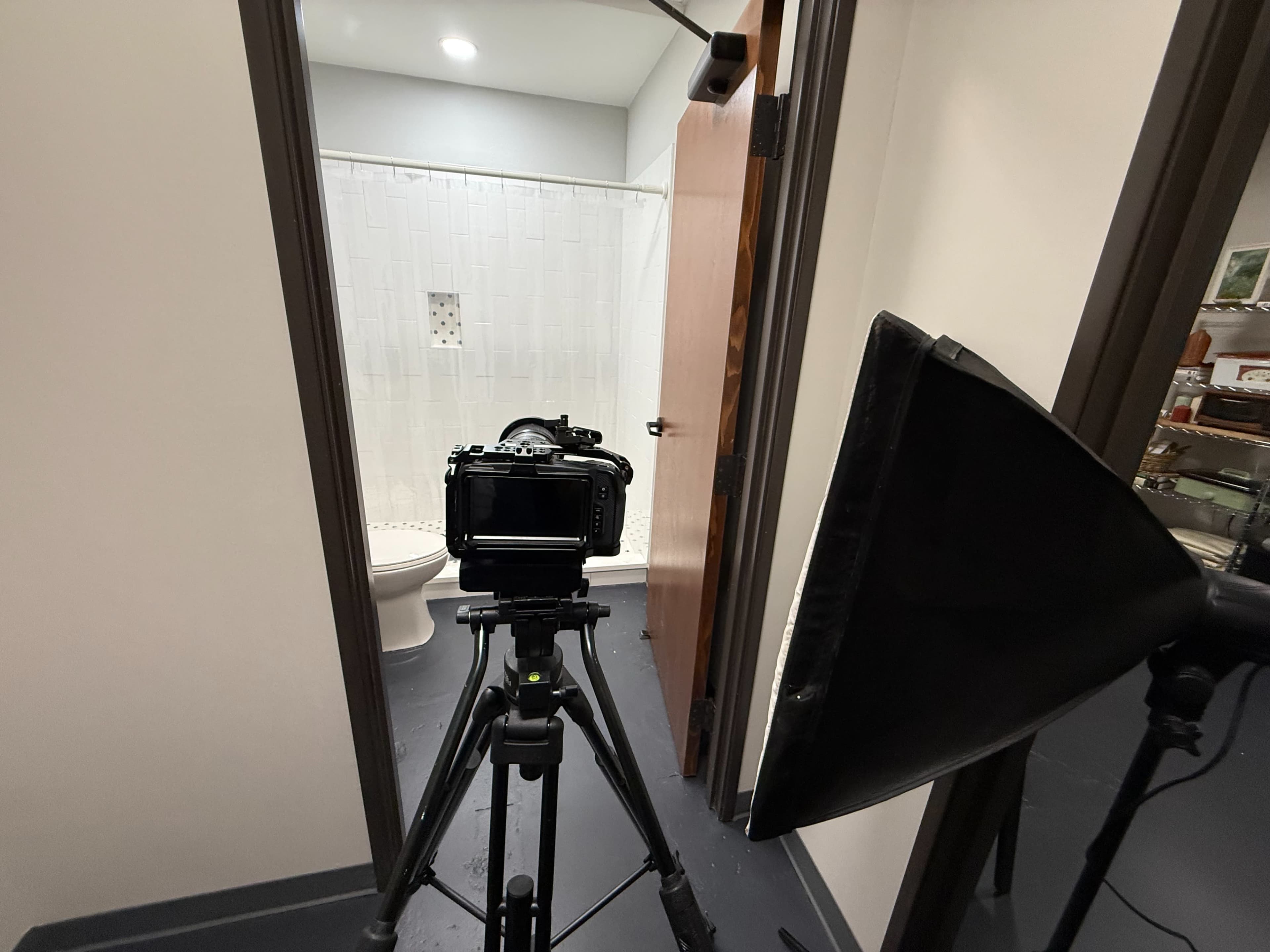 A camera is set on a tripod facing a bathroom doorway, with visible toilet and shower curtain inside.
