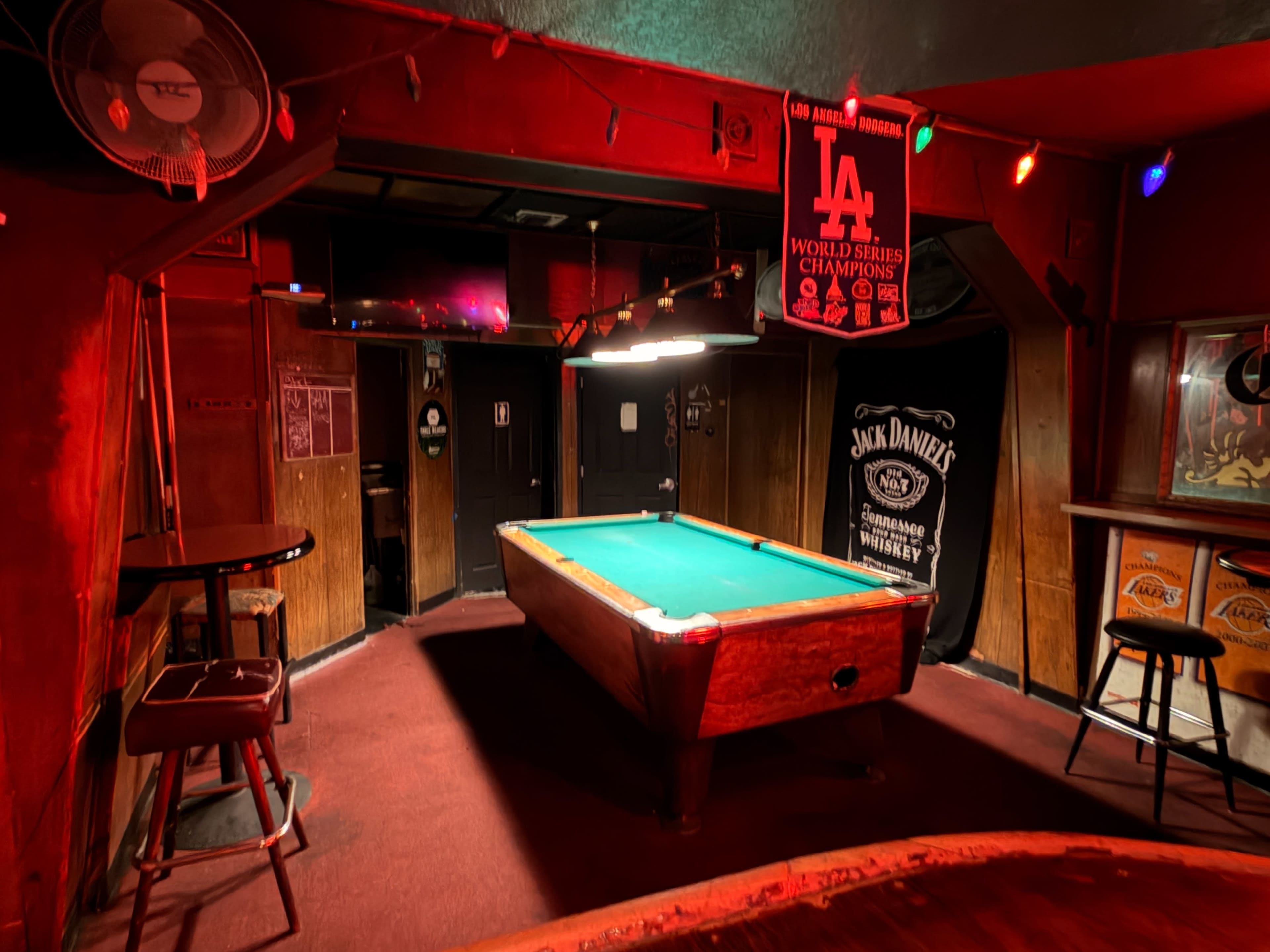 The image shows a dimly lit barroom with a green felt pool table at the center and wooden paneling on the walls.