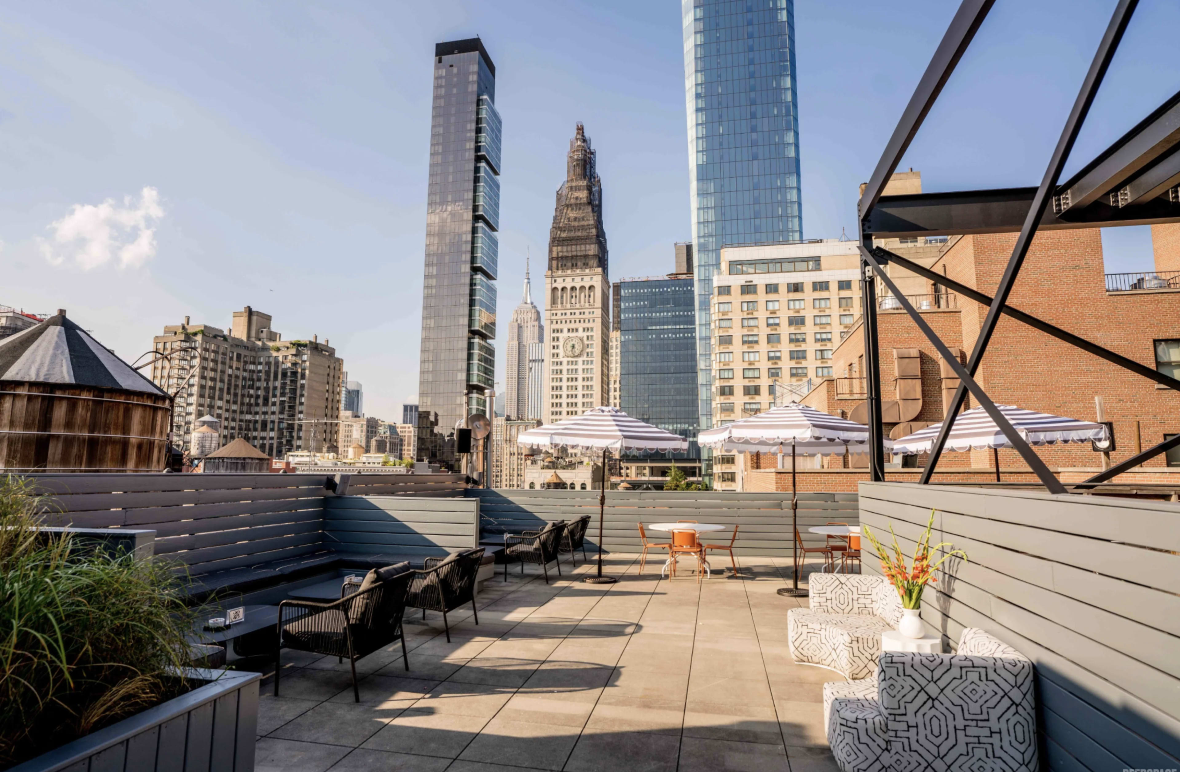The image shows a rooftop terrace with seating areas and umbrellas, overlooking a cityscape featuring tall buildings.