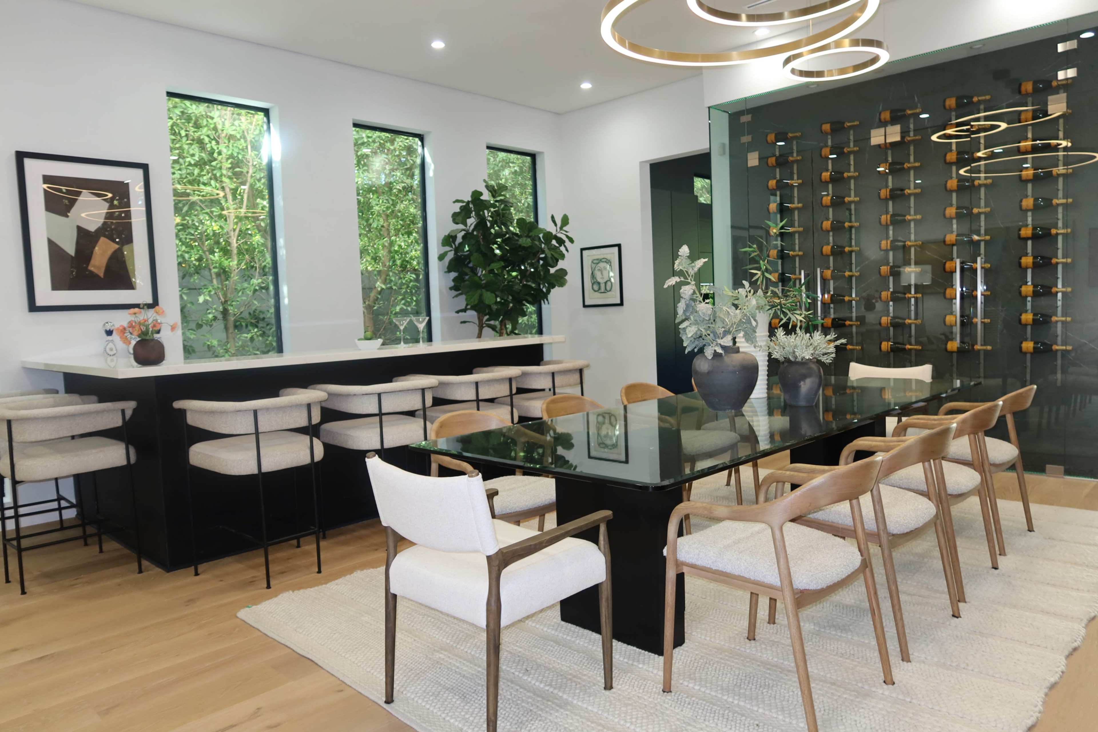 A modern dining area features a black rectangular table surrounded by wooden chairs, a bar counter with stools, large windows allowing natural light, and a wall displaying an assortment of wine bottles.