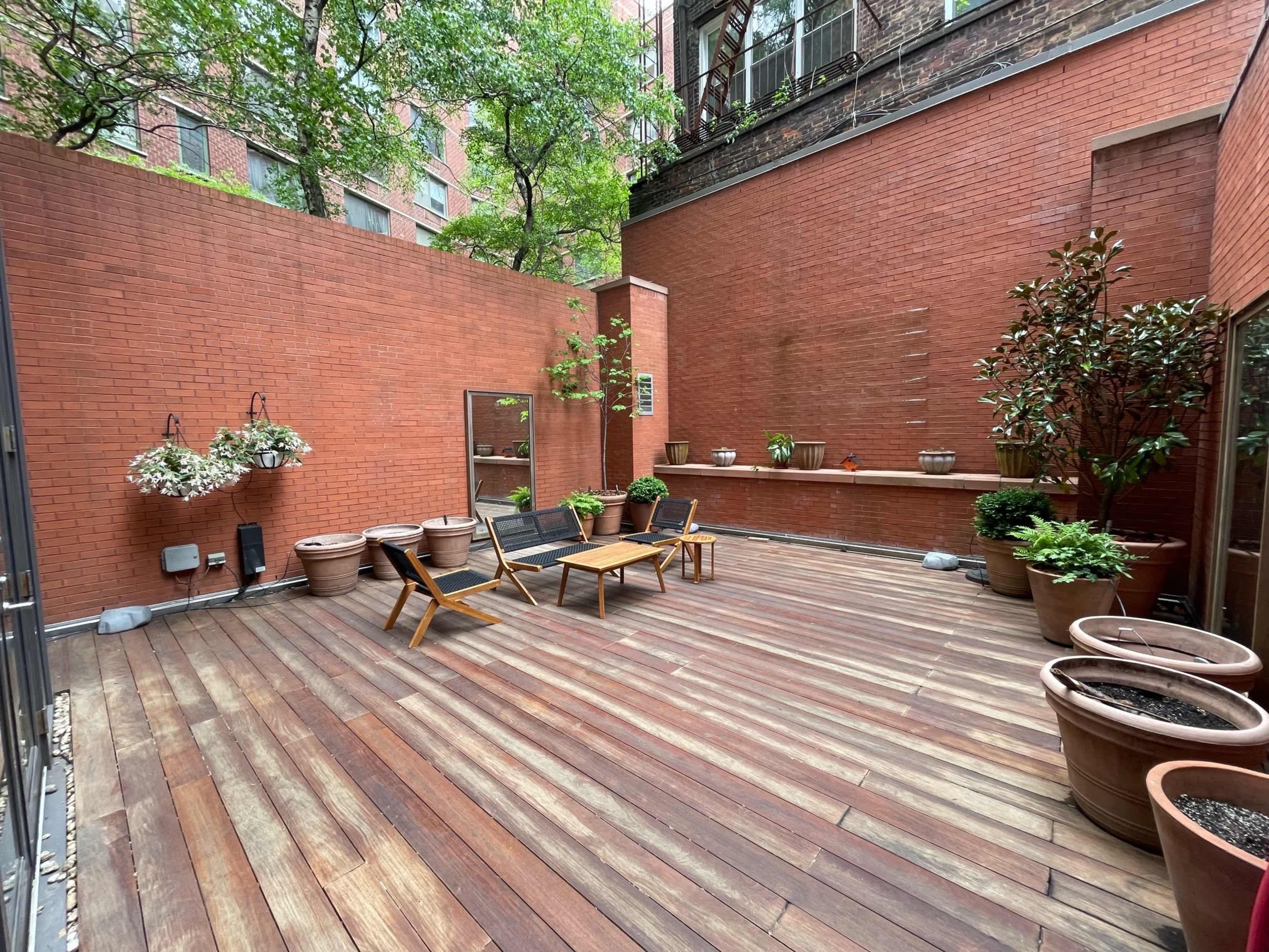 A spacious outdoor patio with wooden flooring, potted plants, and a few wooden chairs and tables.