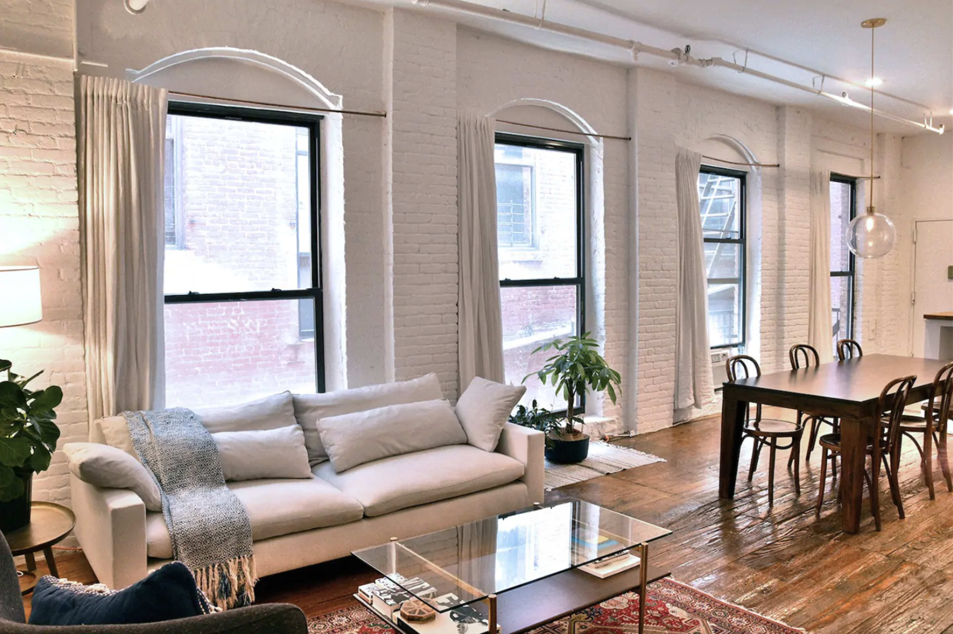 A cozy living space with a white couch, a wooden dining table, and large windows letting in natural light.