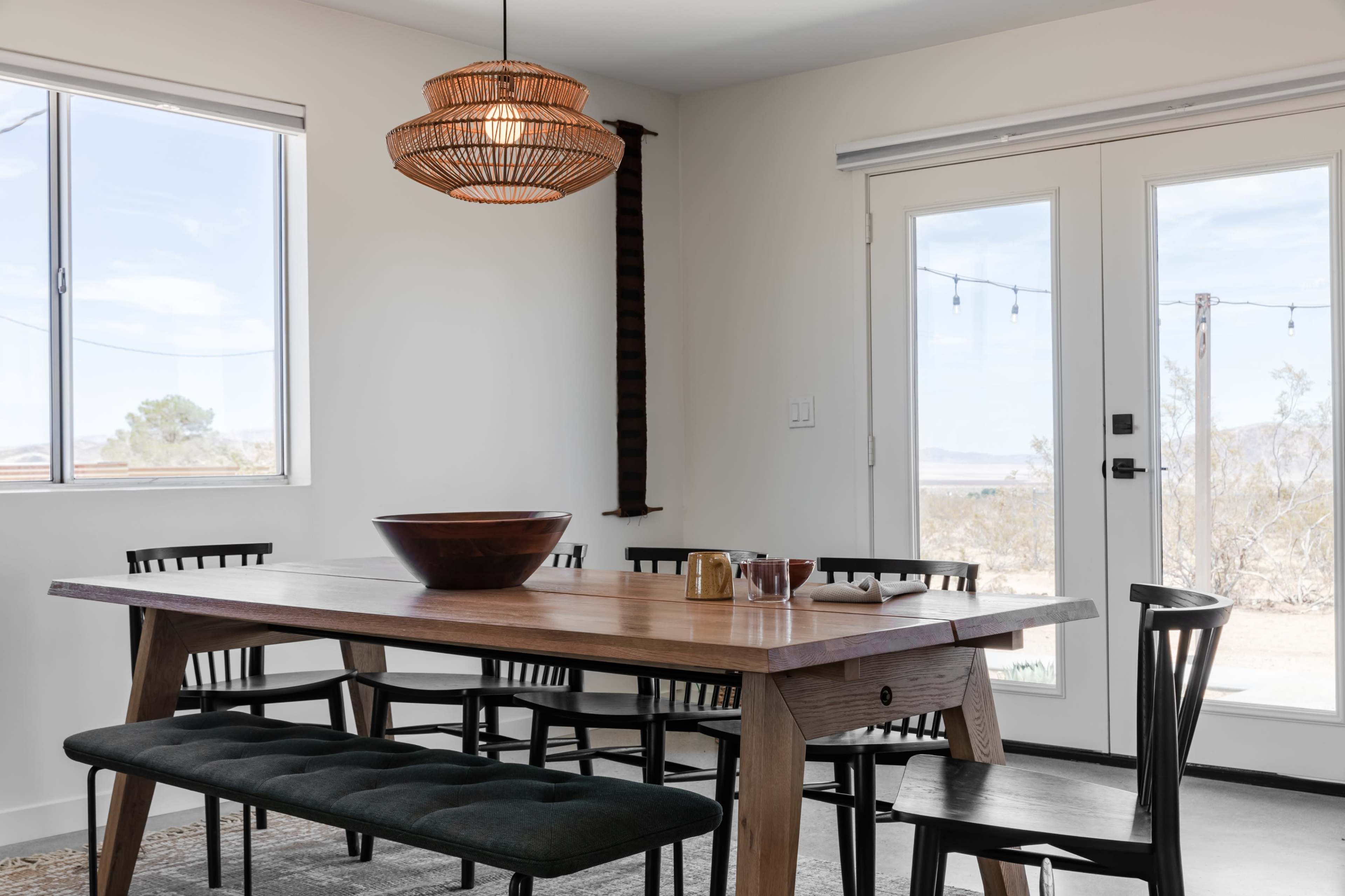 A wooden dining table with black chairs and a bench is set in a light-filled room featuring large windows and double doors leading to an outdoor area.