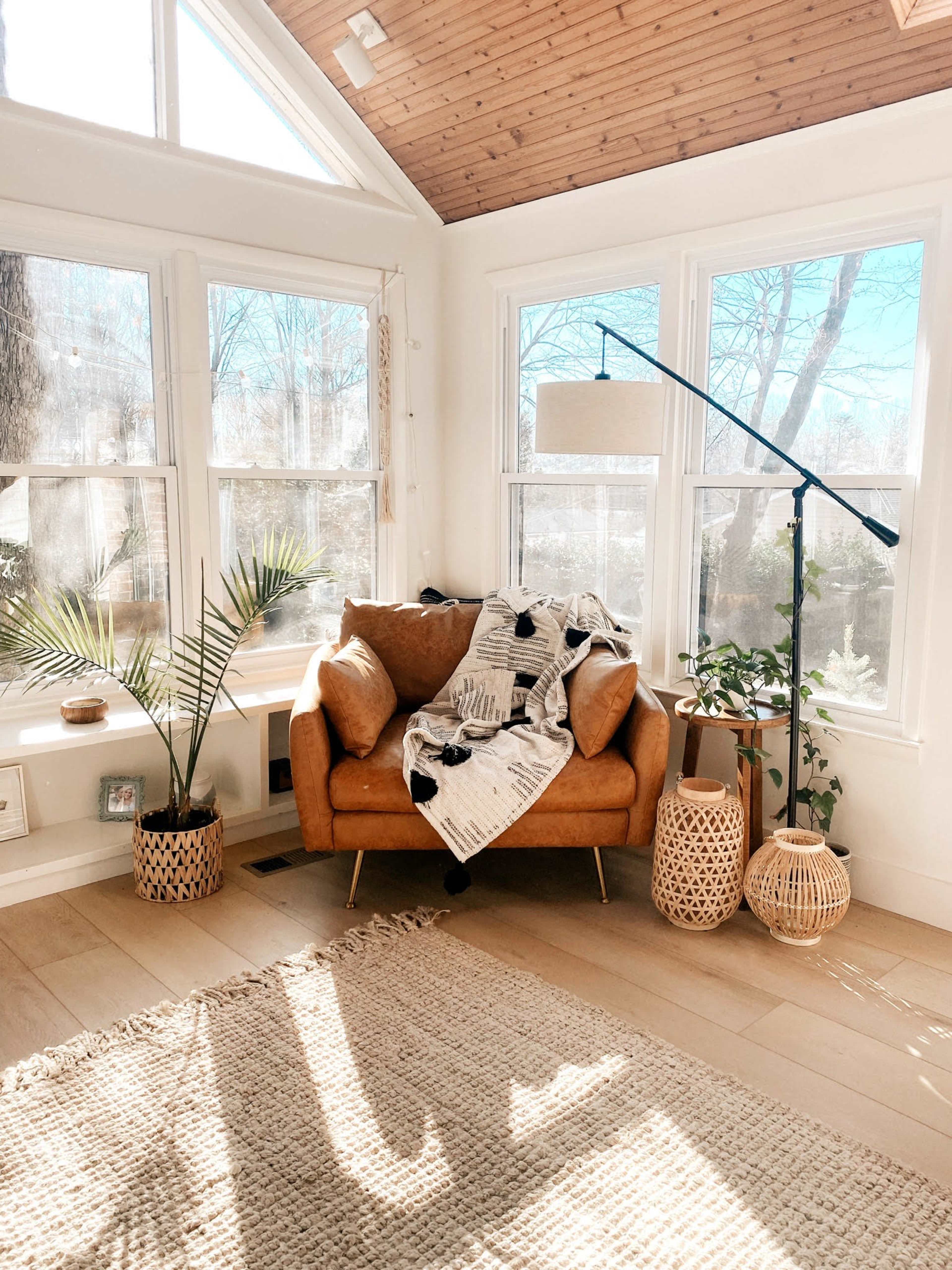 The image shows a cozy living space with a brown leather sofa, a patterned blanket draped over it, houseplants beside it, and large windows allowing natural light to fill the room.