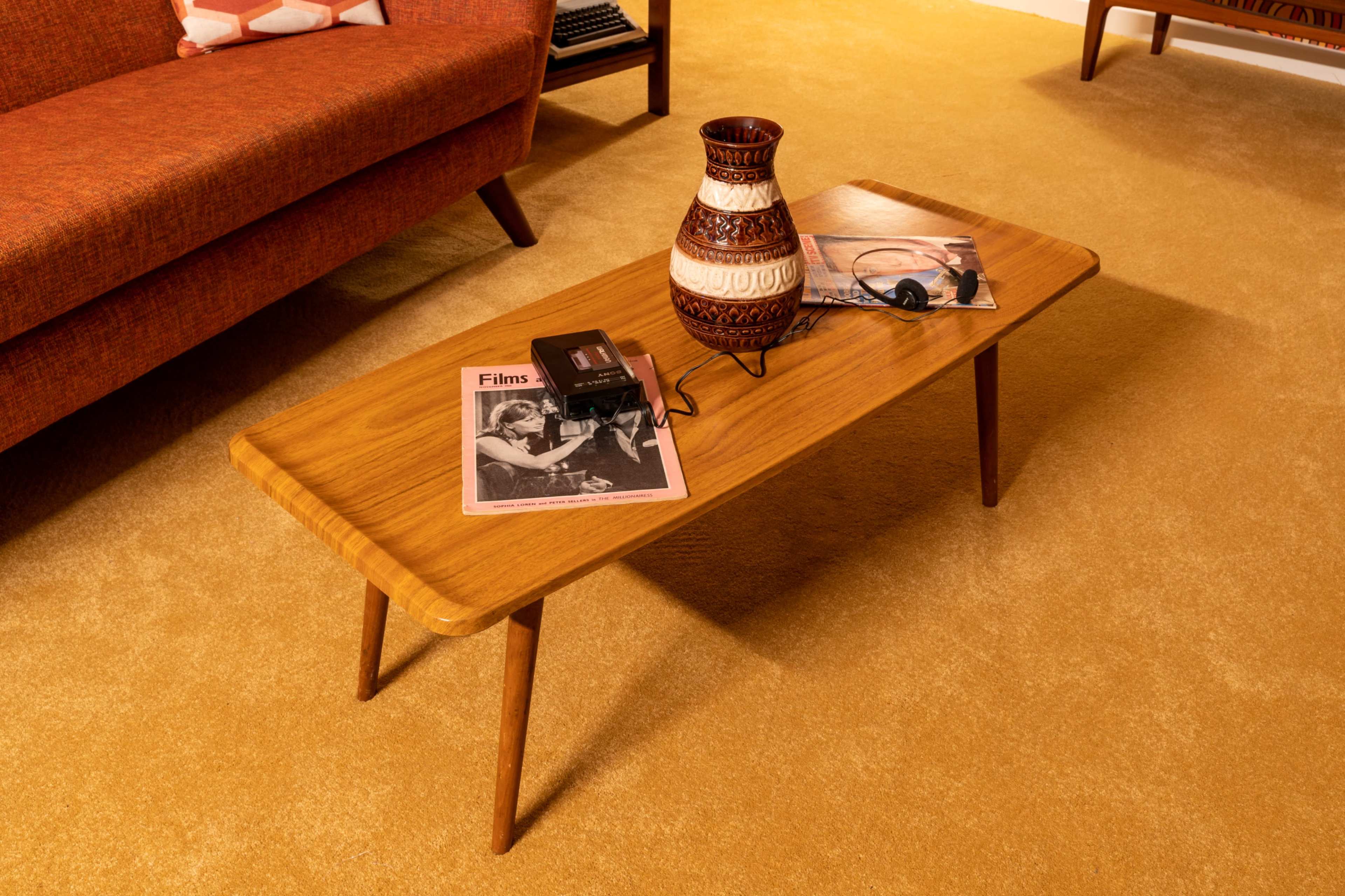 A wooden coffee table with a vase and magazines sits on a carpeted floor in a retro-styled living room.