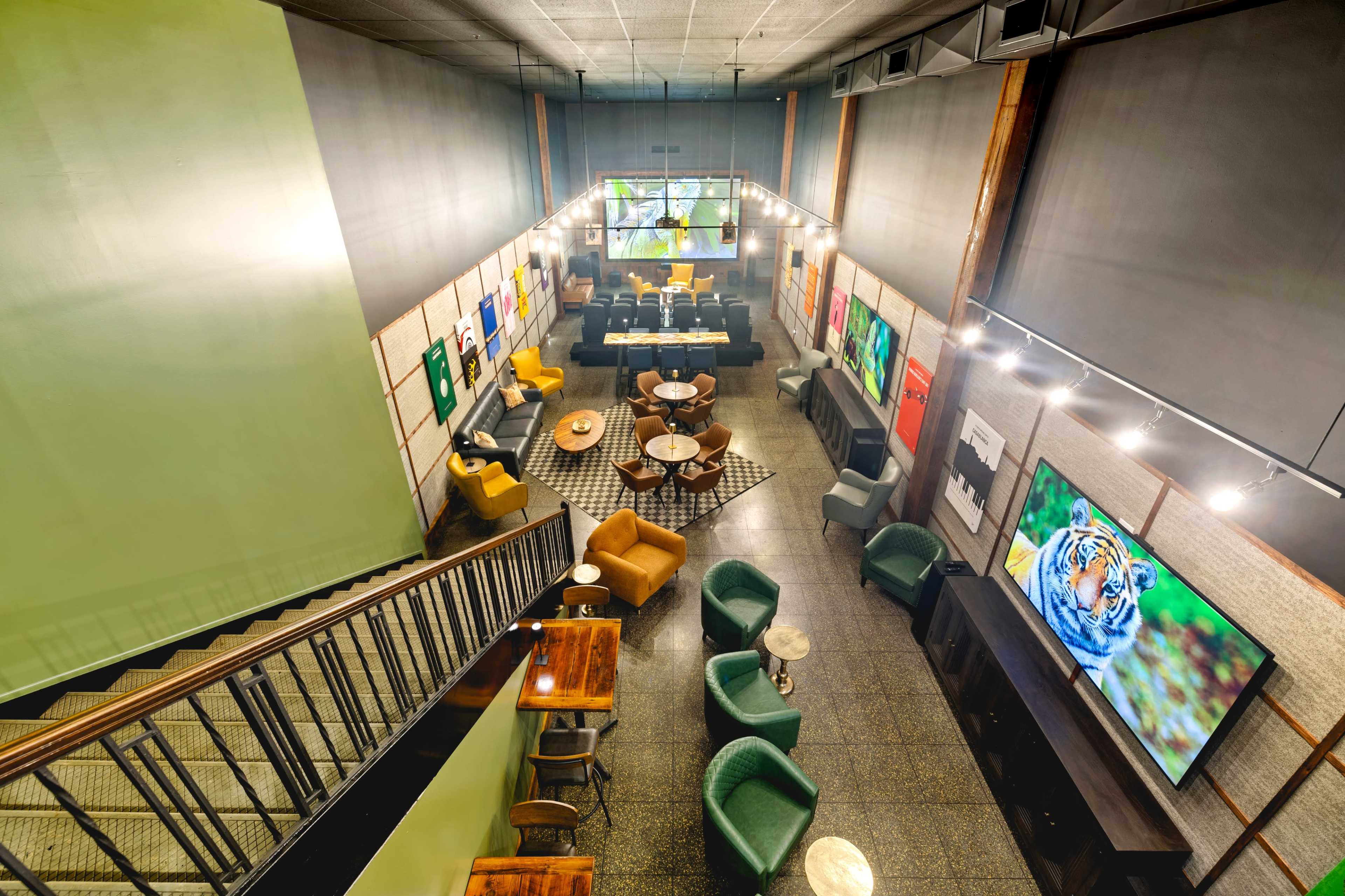 The image shows an interior space with a modern café or lounge setup, featuring a variety of seating arrangements, artworks on the walls, and large screens displaying images.