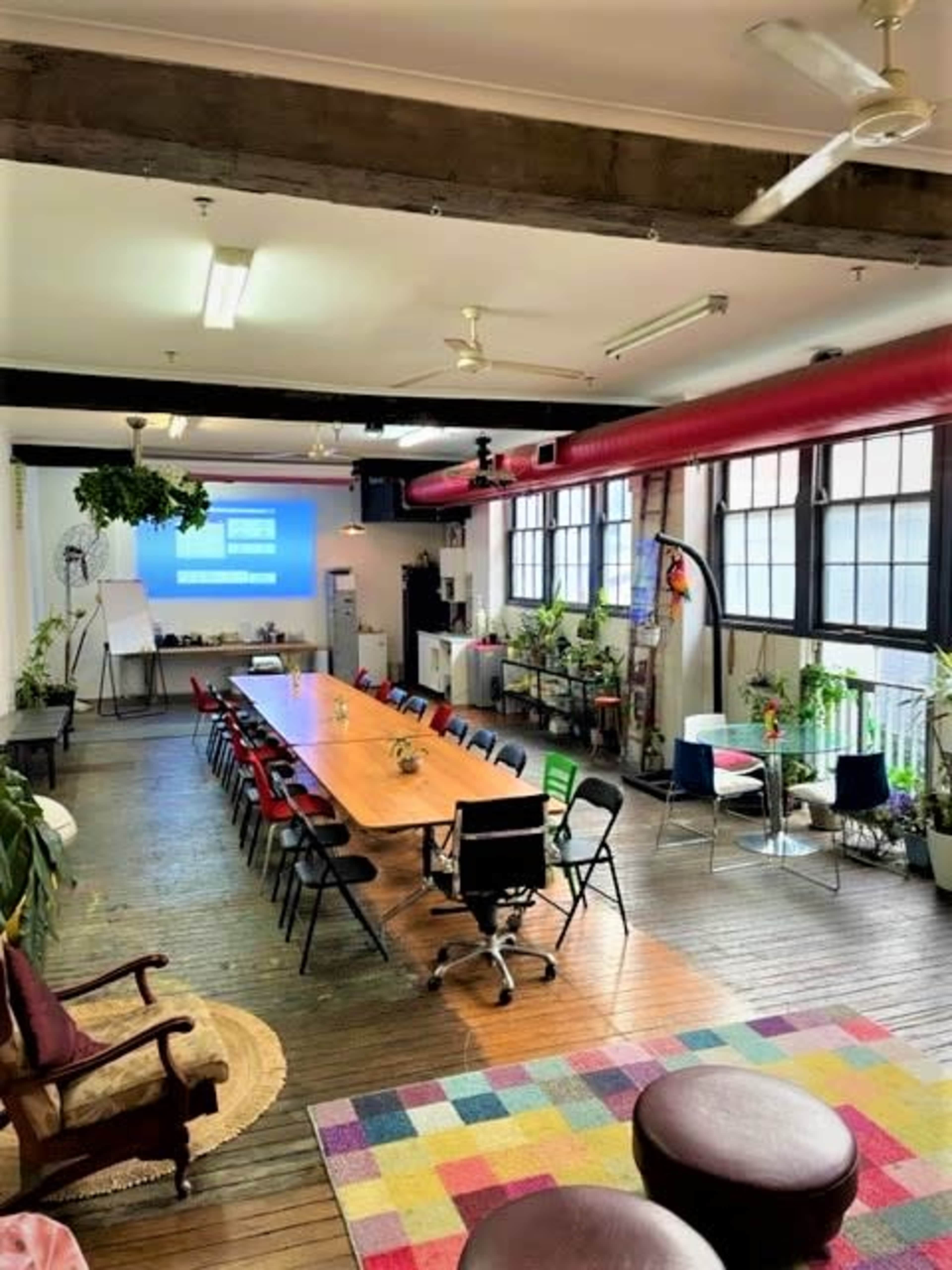 A spacious meeting room features a long wooden table surrounded by chairs, with large windows allowing natural light and a colorful rug on the floor.