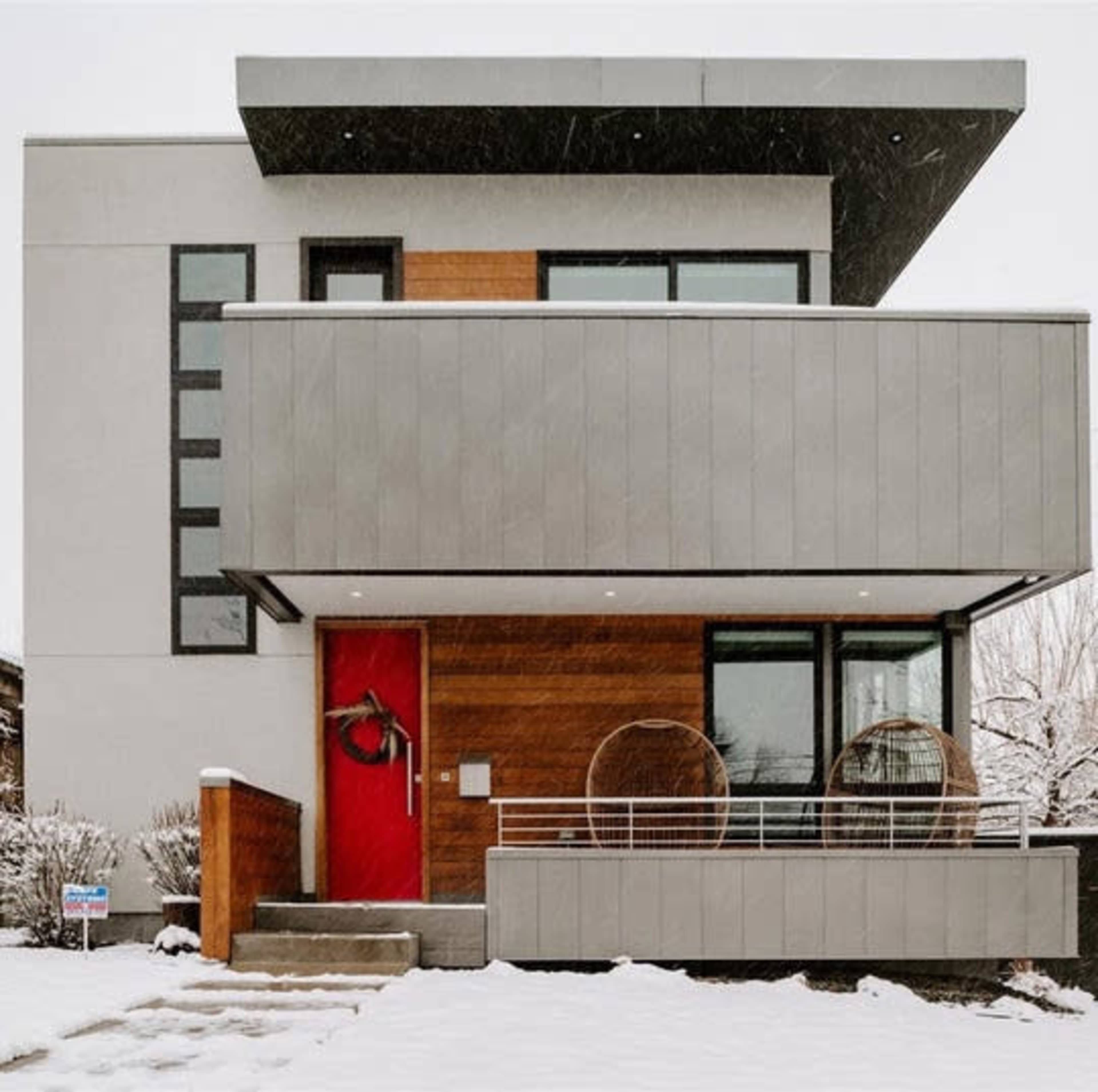 A modern house with a red front door, featuring a balcony and surrounded by snow.