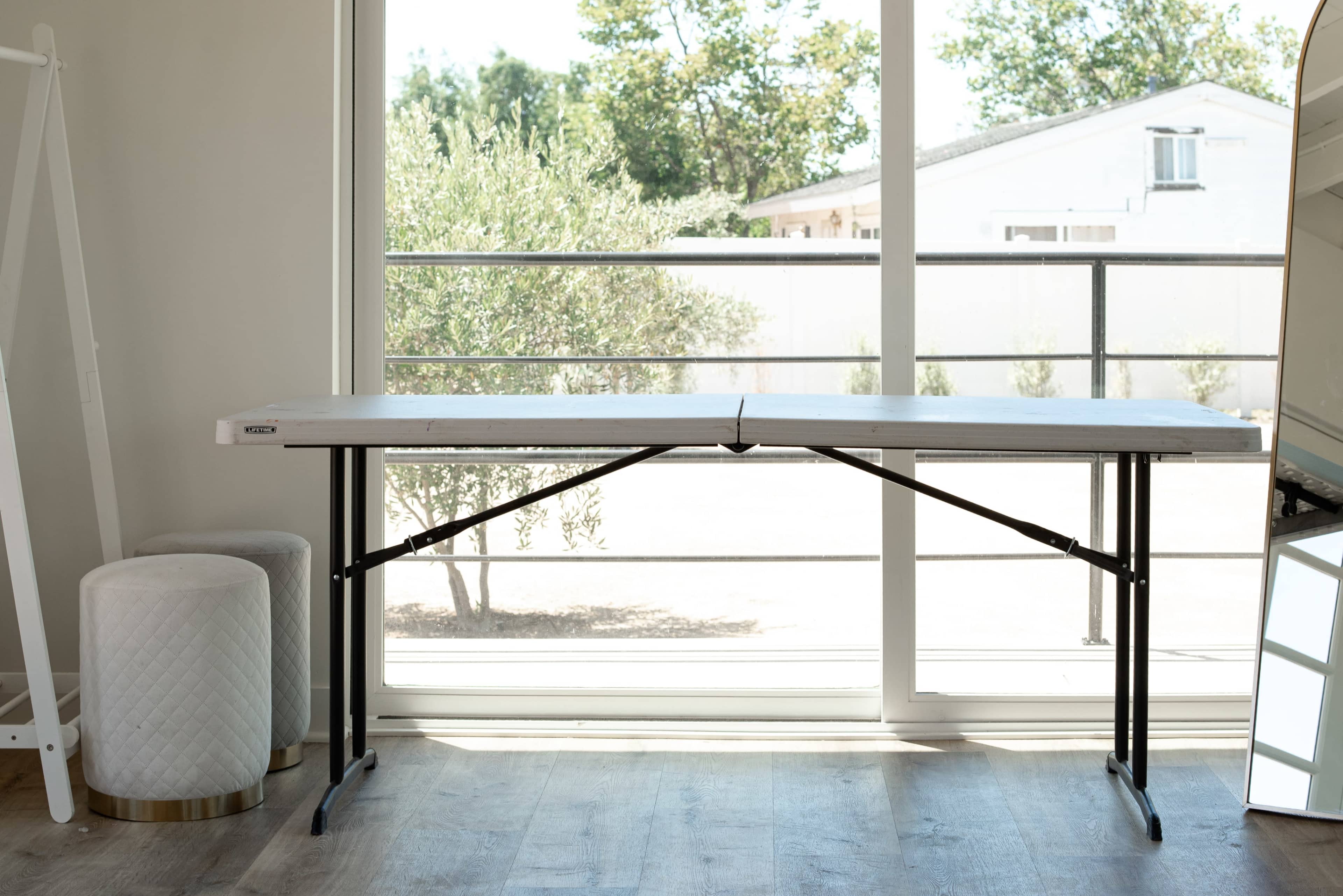 A foldable table is positioned in front of a large window that lets in natural light, with a mirror and two round stools nearby.
