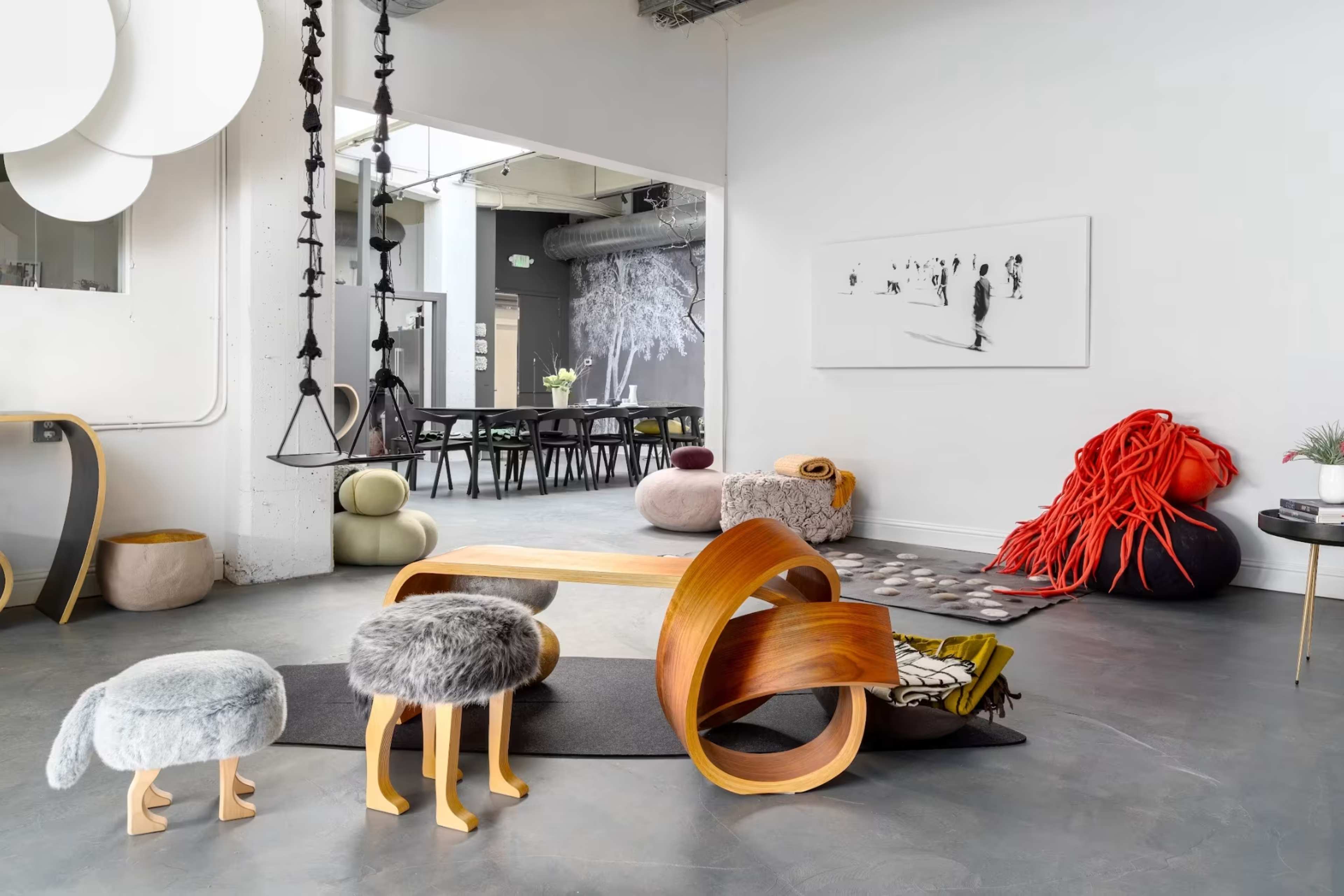 A modern interior space featuring various furniture pieces, including a wooden table, uniquely designed stools, and textured poufs, decorated with art on the walls.