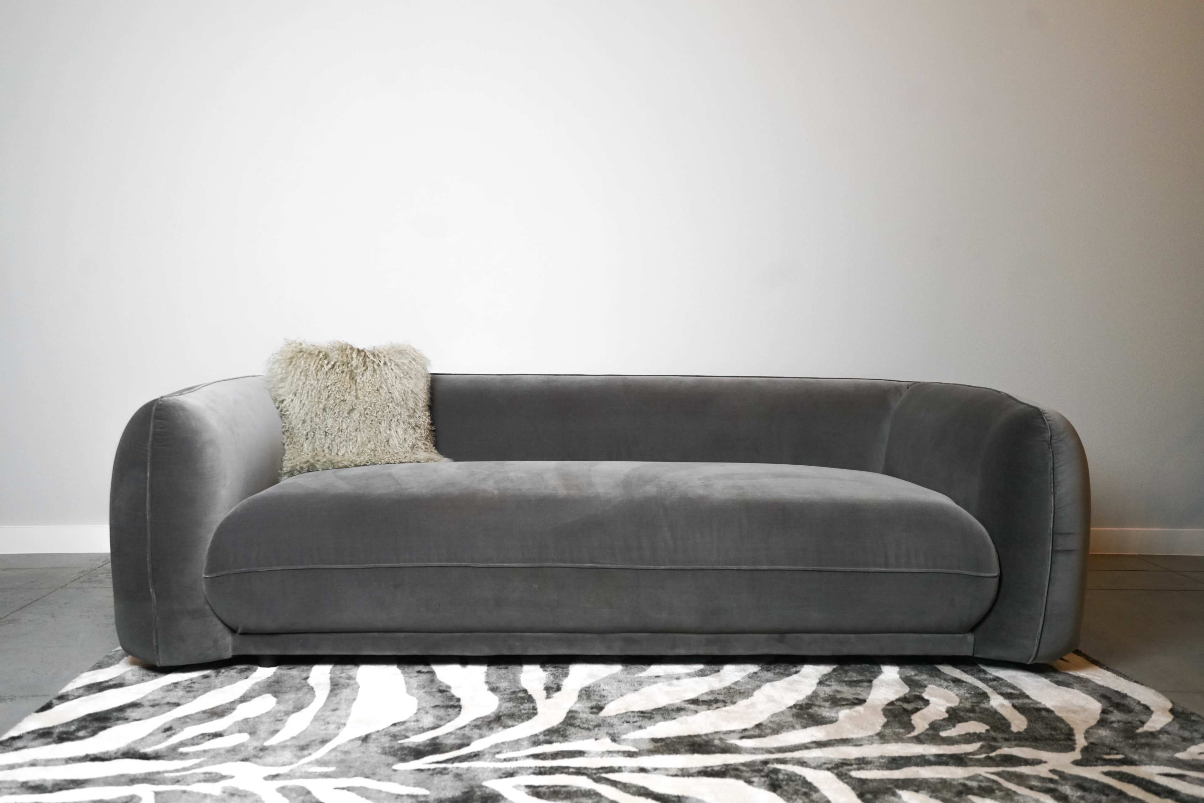A gray upholstered sofa with rounded arms and a textured pillow sits on a zebra-striped rug against a plain wall.