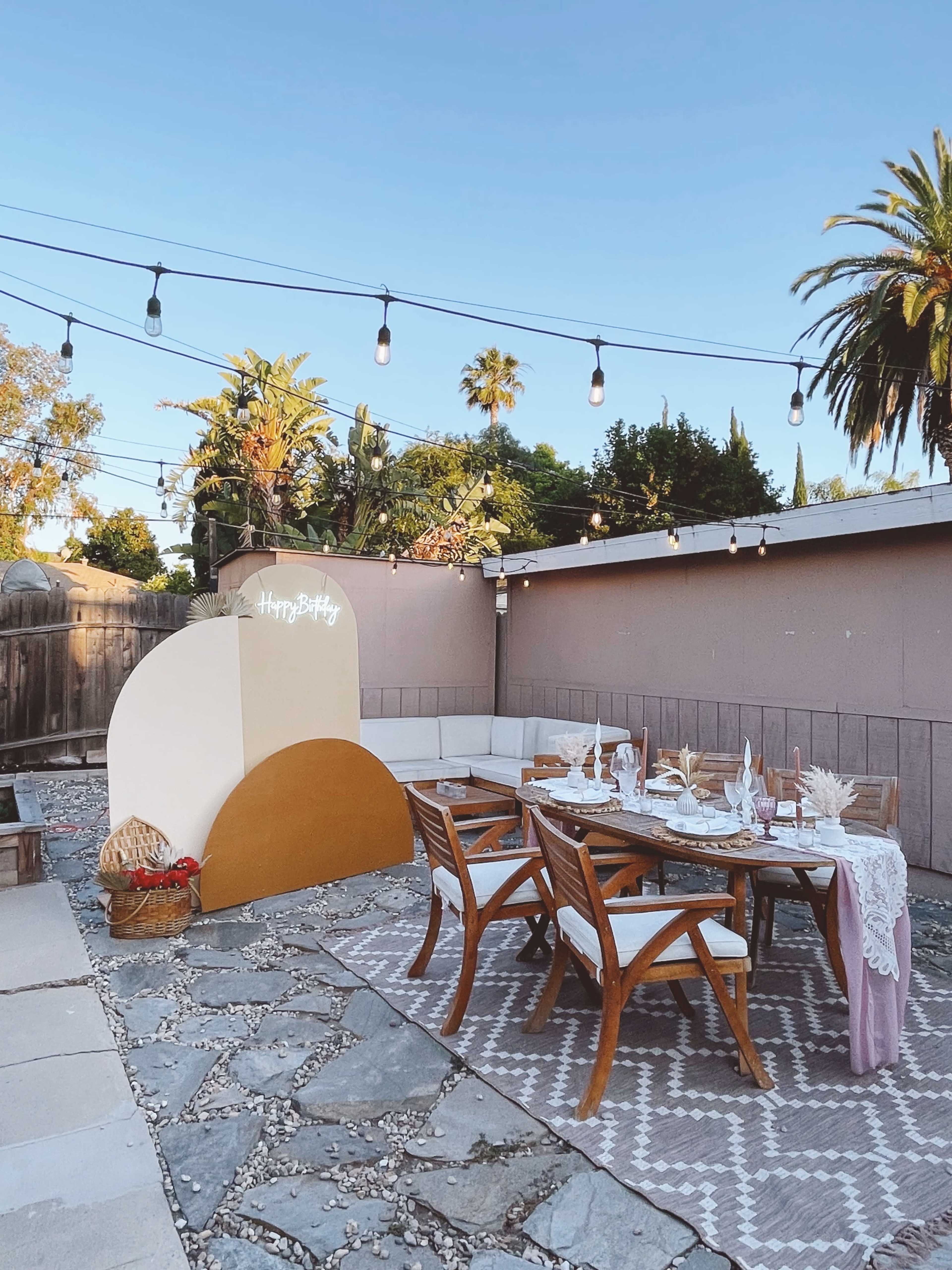 A decorated outdoor dining area features a wooden table set with plates and glasses, surrounded by a stone patio and palm trees, with string lights overhead.