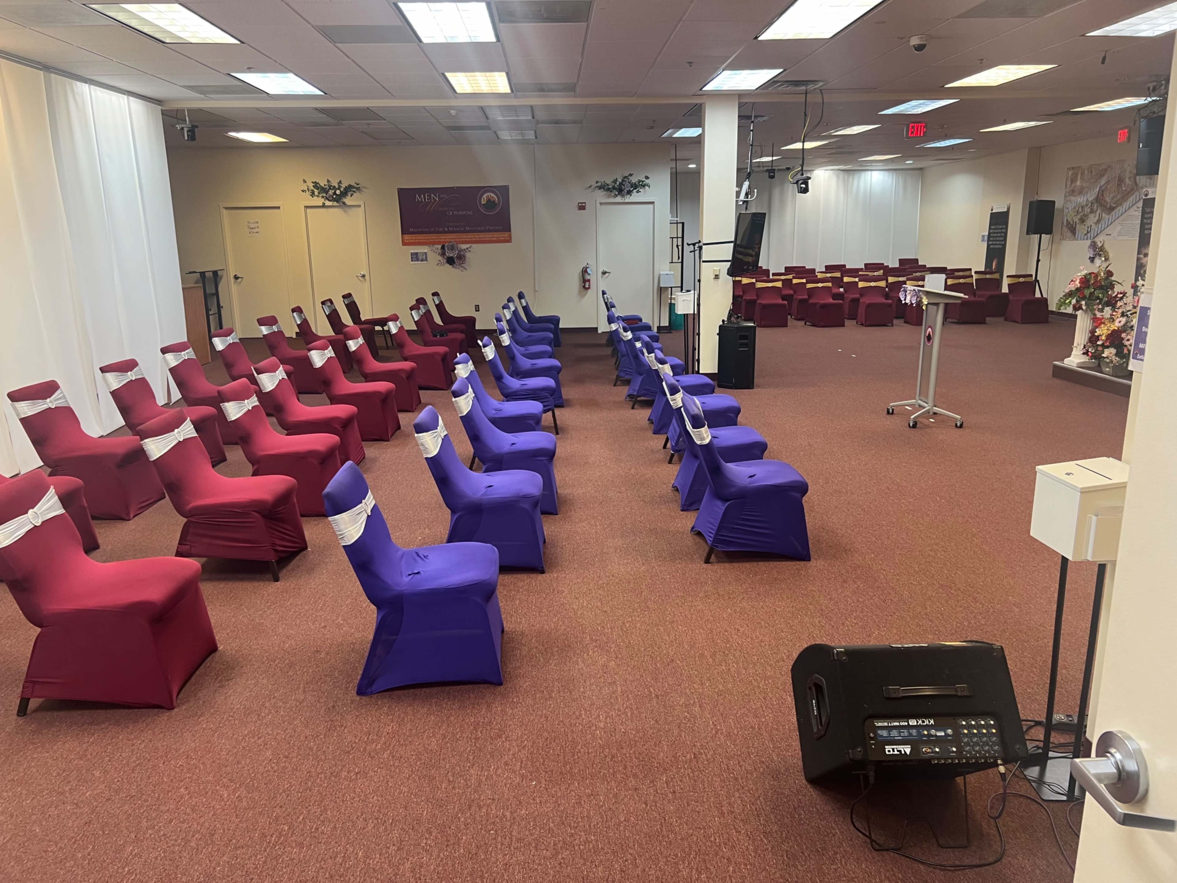 The image shows a large room set up for an event, with rows of chairs covered in red and purple fabric, and a presentation area at the front.