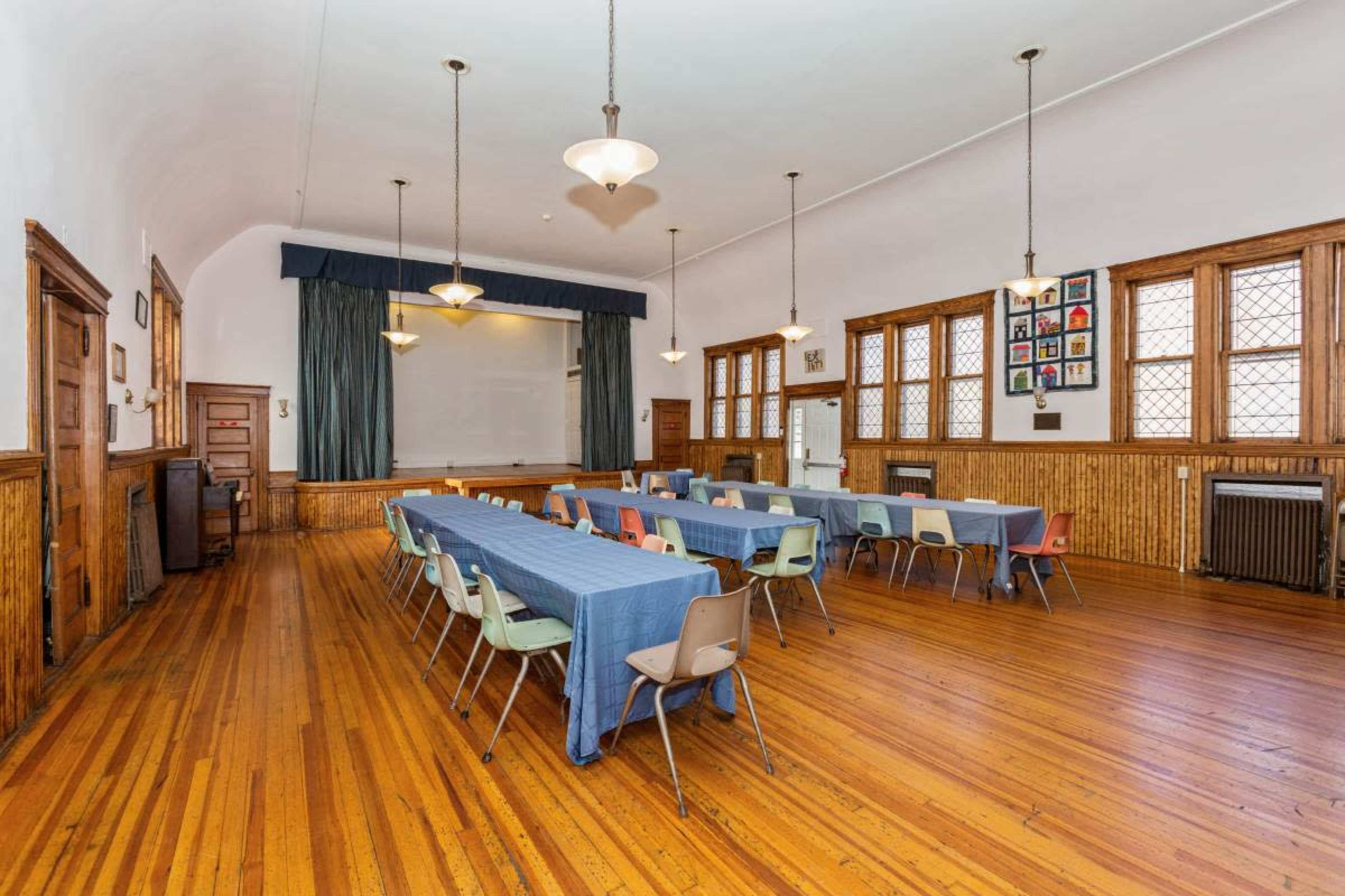 A large, empty room features a long table set with chairs, hardwood floors, and windows with decorative panes.