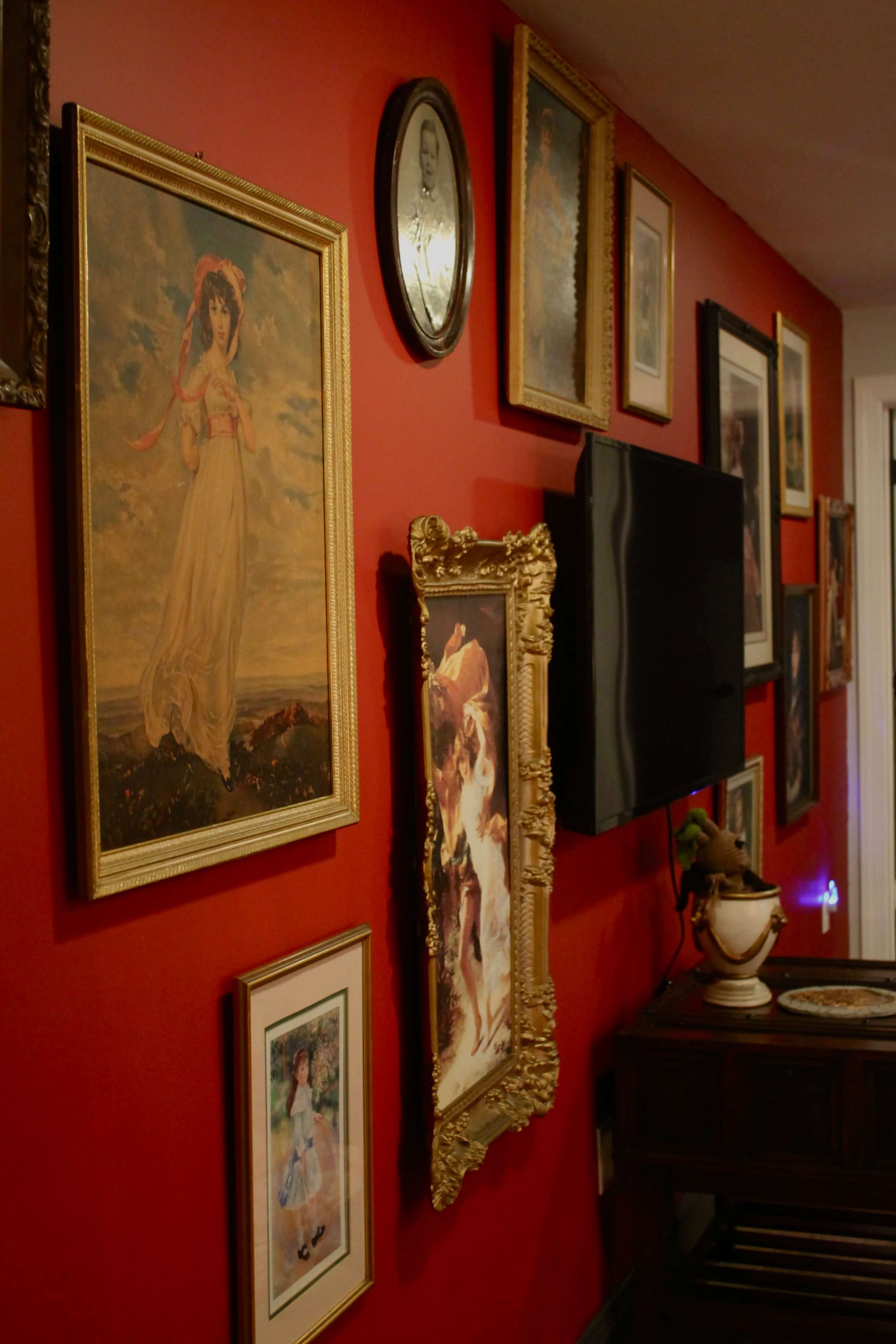 The image shows a wall adorned with various framed artworks and photographs against a red background, with a television mounted in the center.