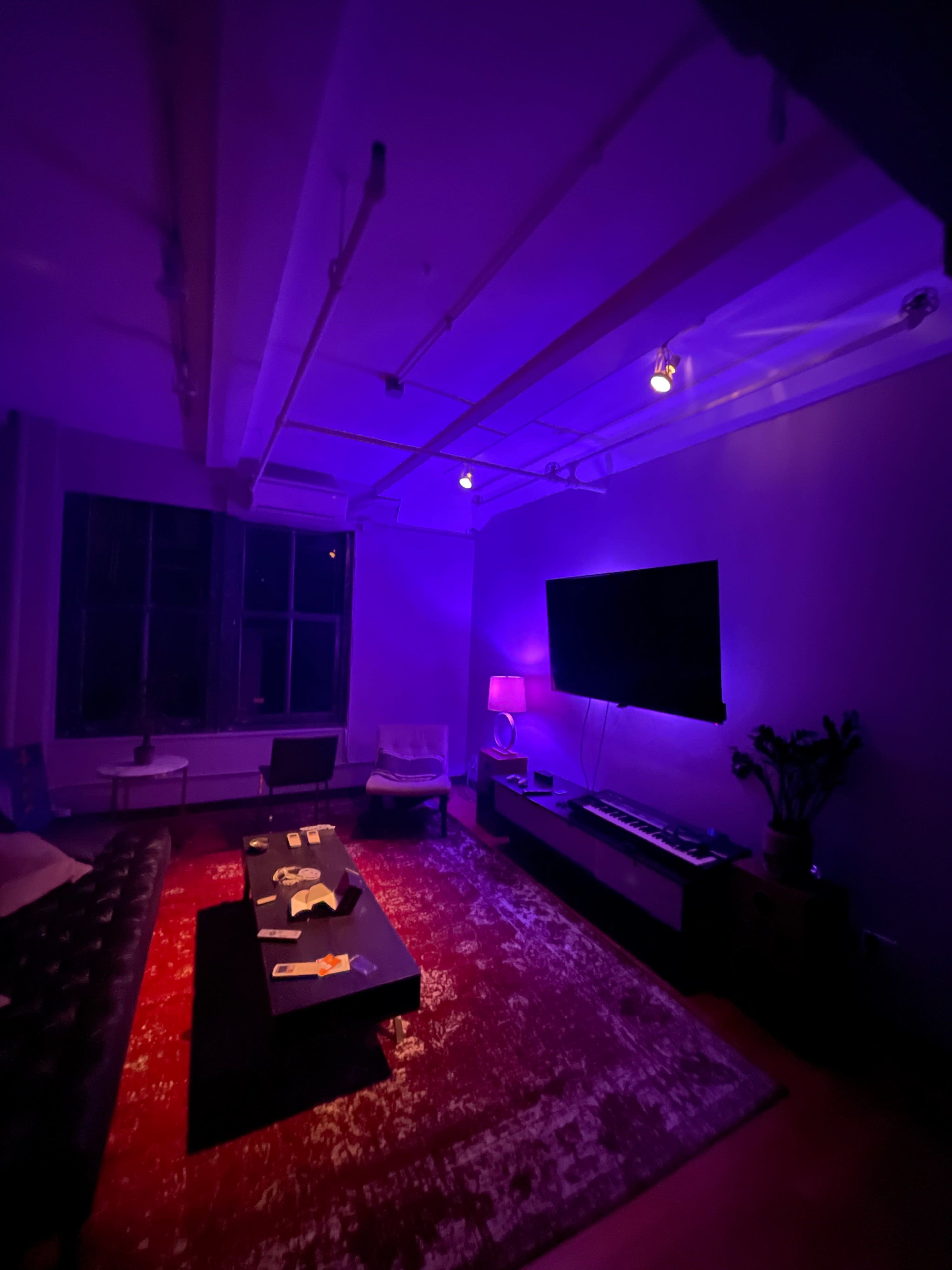 A dimly lit living room features purple lighting, a large television, a coffee table with items on it, and a chair beside a window.
