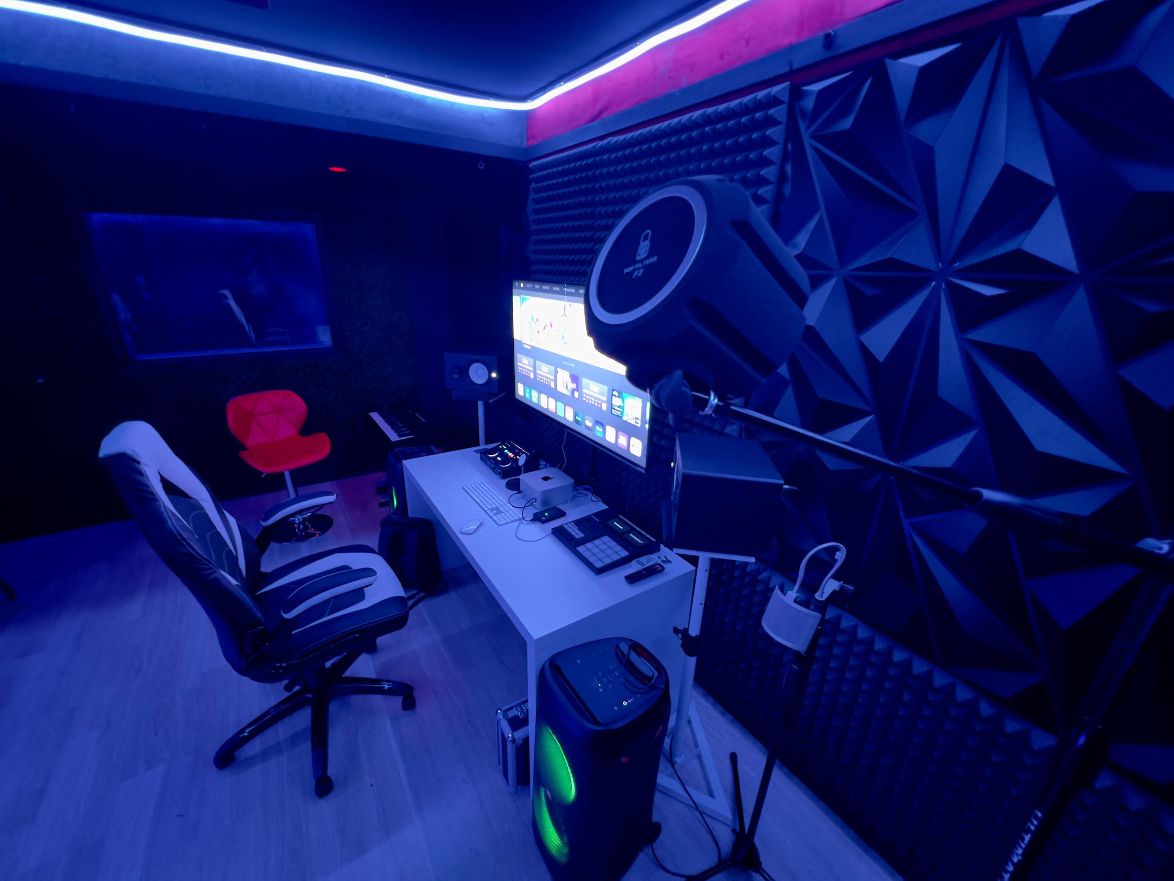 A modern music studio with a white desk, a comfortable chair, and various audio equipment, illuminated by blue LED lights.