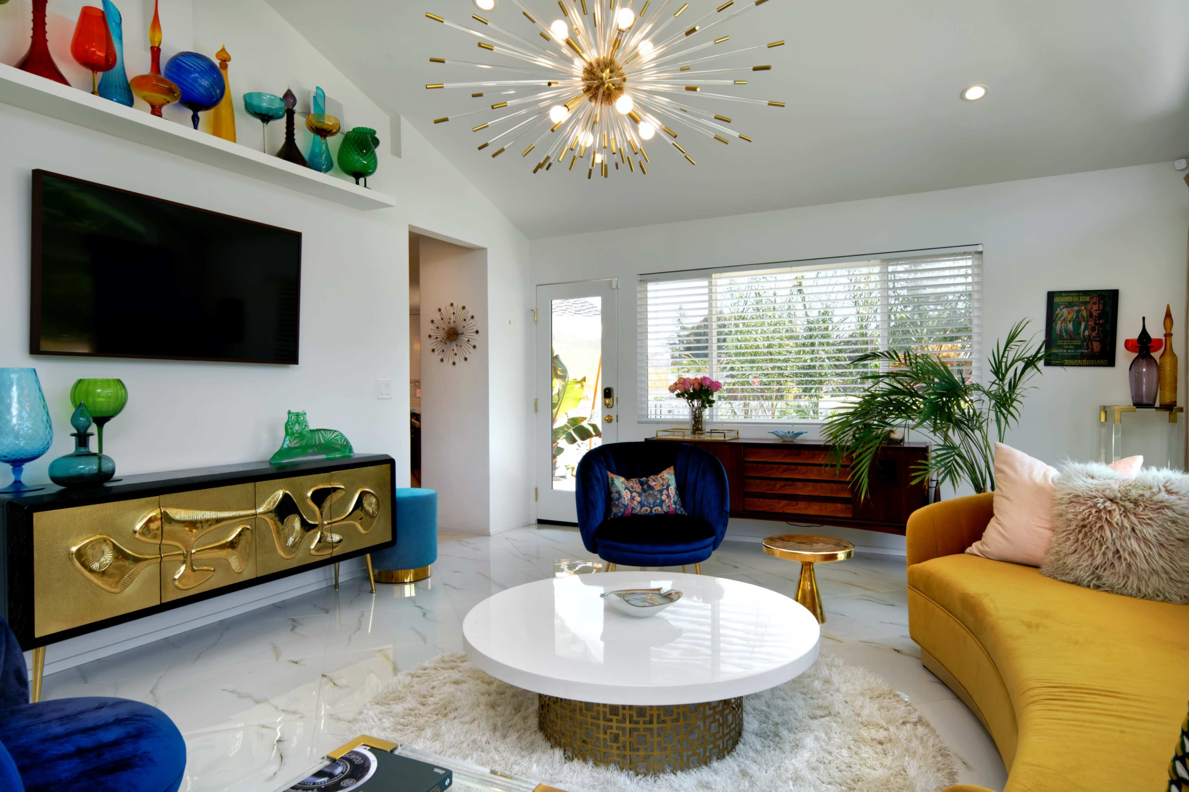 The image shows a modern living room featuring a round white coffee table, a yellow and blue velvet sofa, a television, decorative vases, and large windows with natural light coming in.