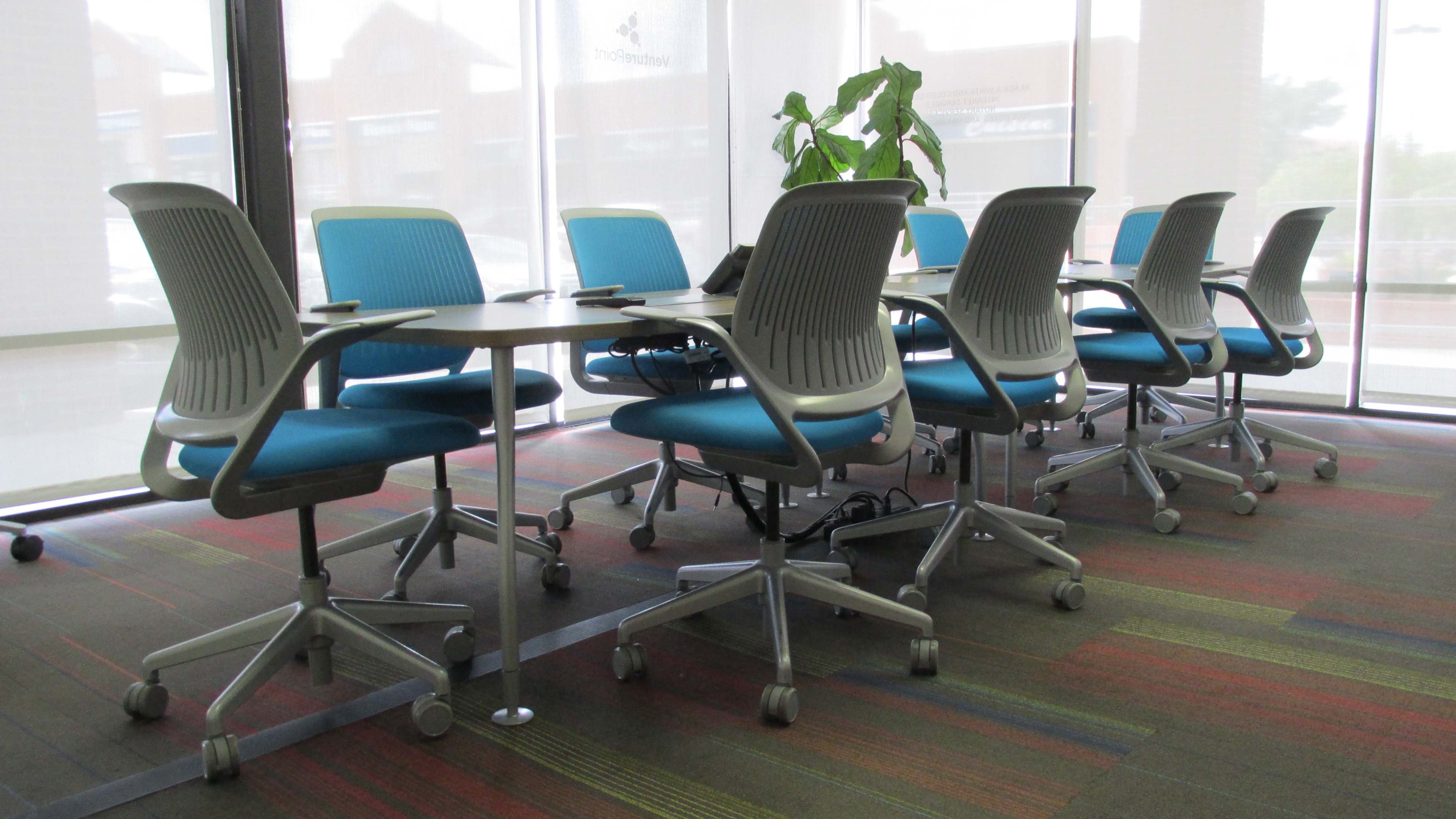 A row of equipped blue chairs and tables is arranged in a brightly lit office space with large windows.