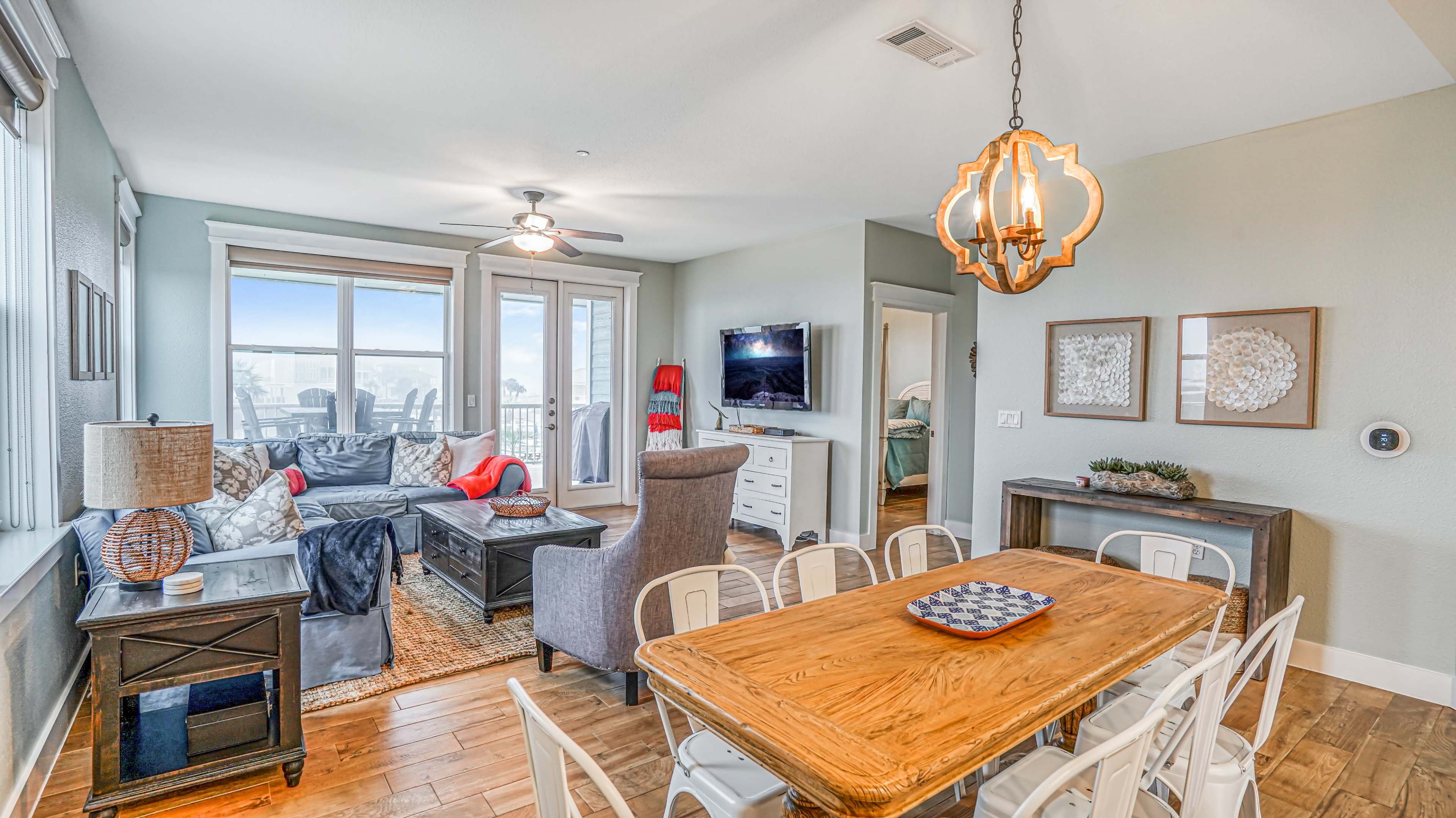 A living and dining area featuring a wooden table with white chairs, a gray couch, and large windows allowing natural light.