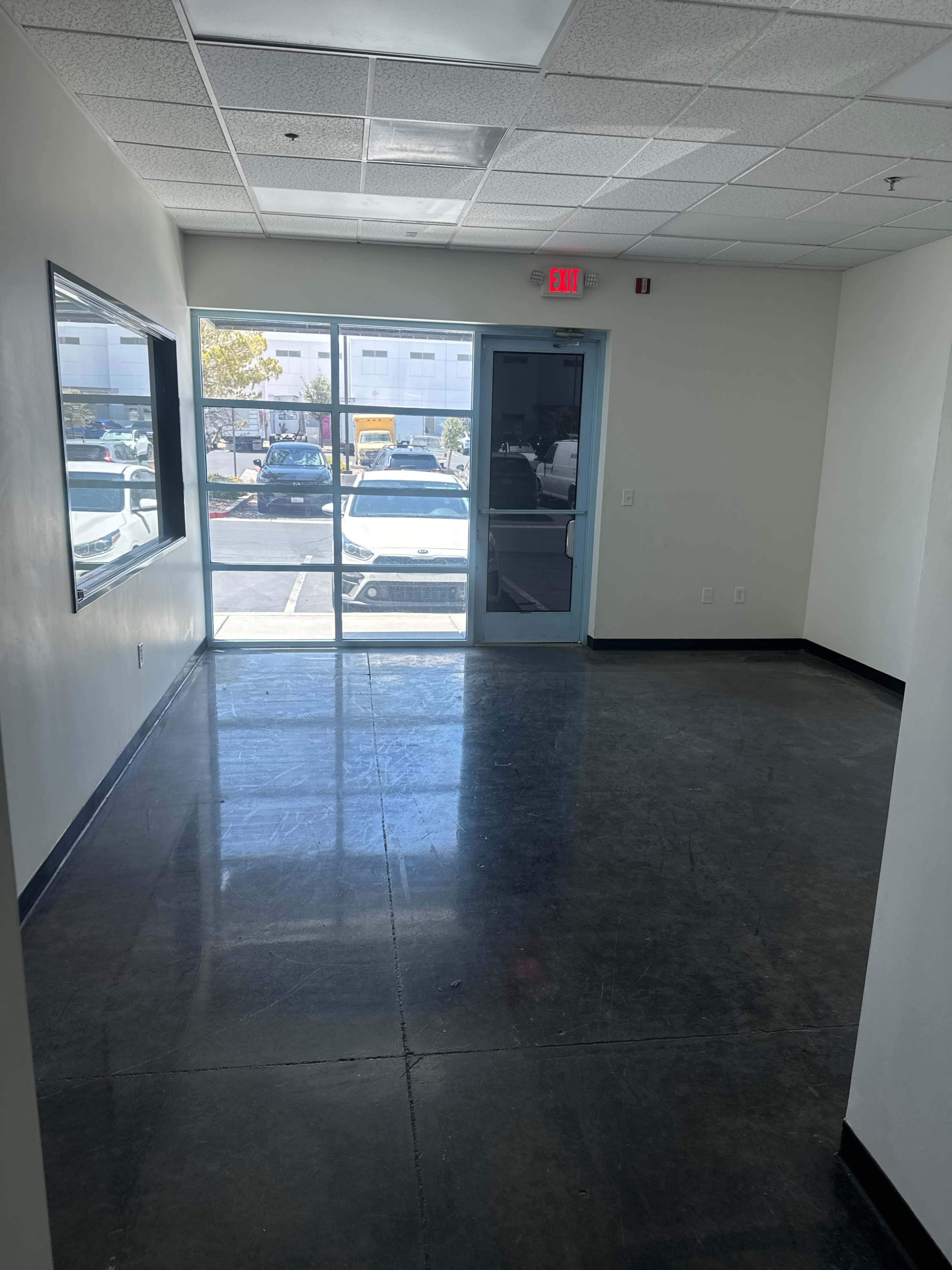 The image shows an empty office space with a large window and a glass door leading to a parking area.