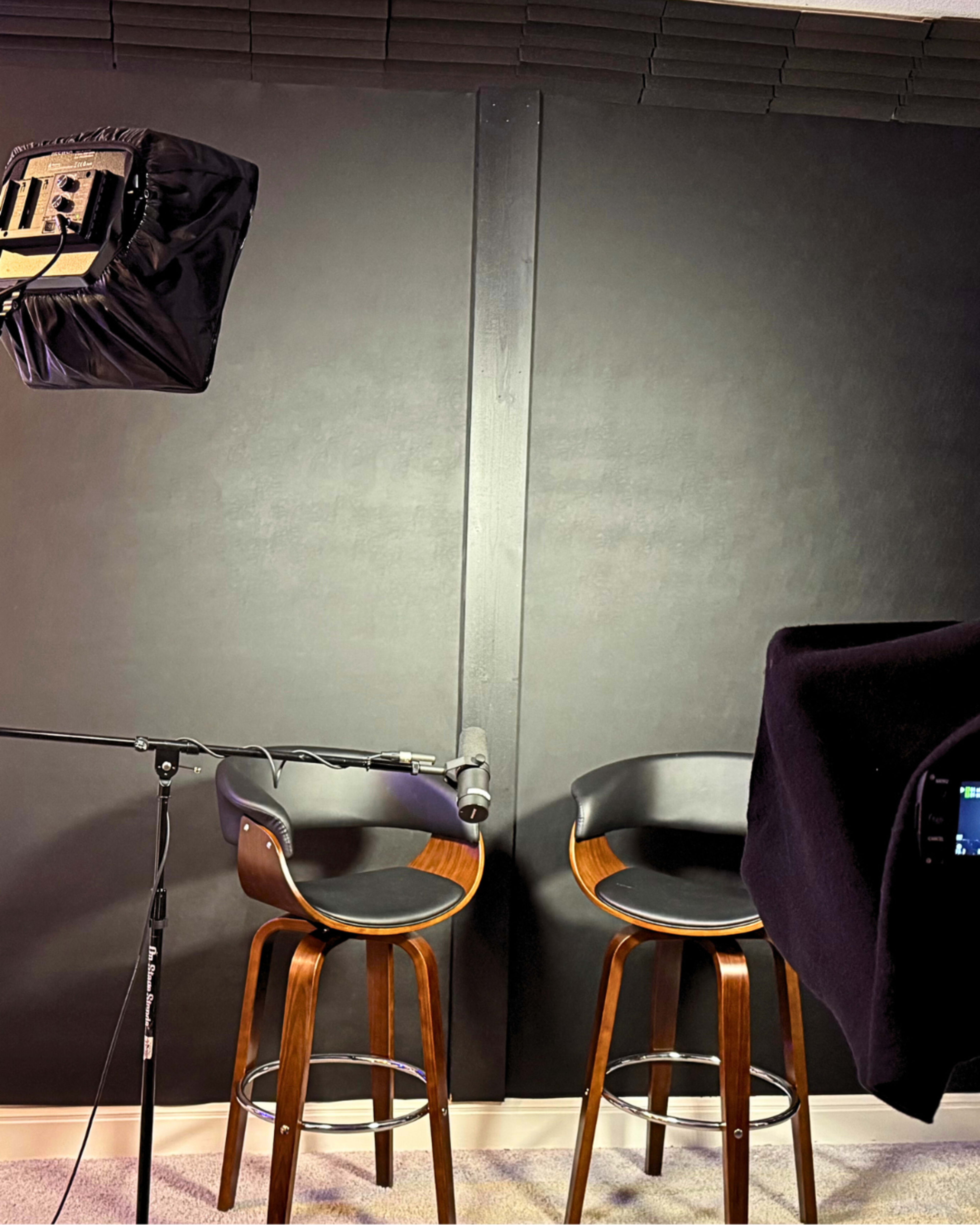 The image shows two bar stools positioned in front of a black wall, with a microphone and a lighting setup in view.