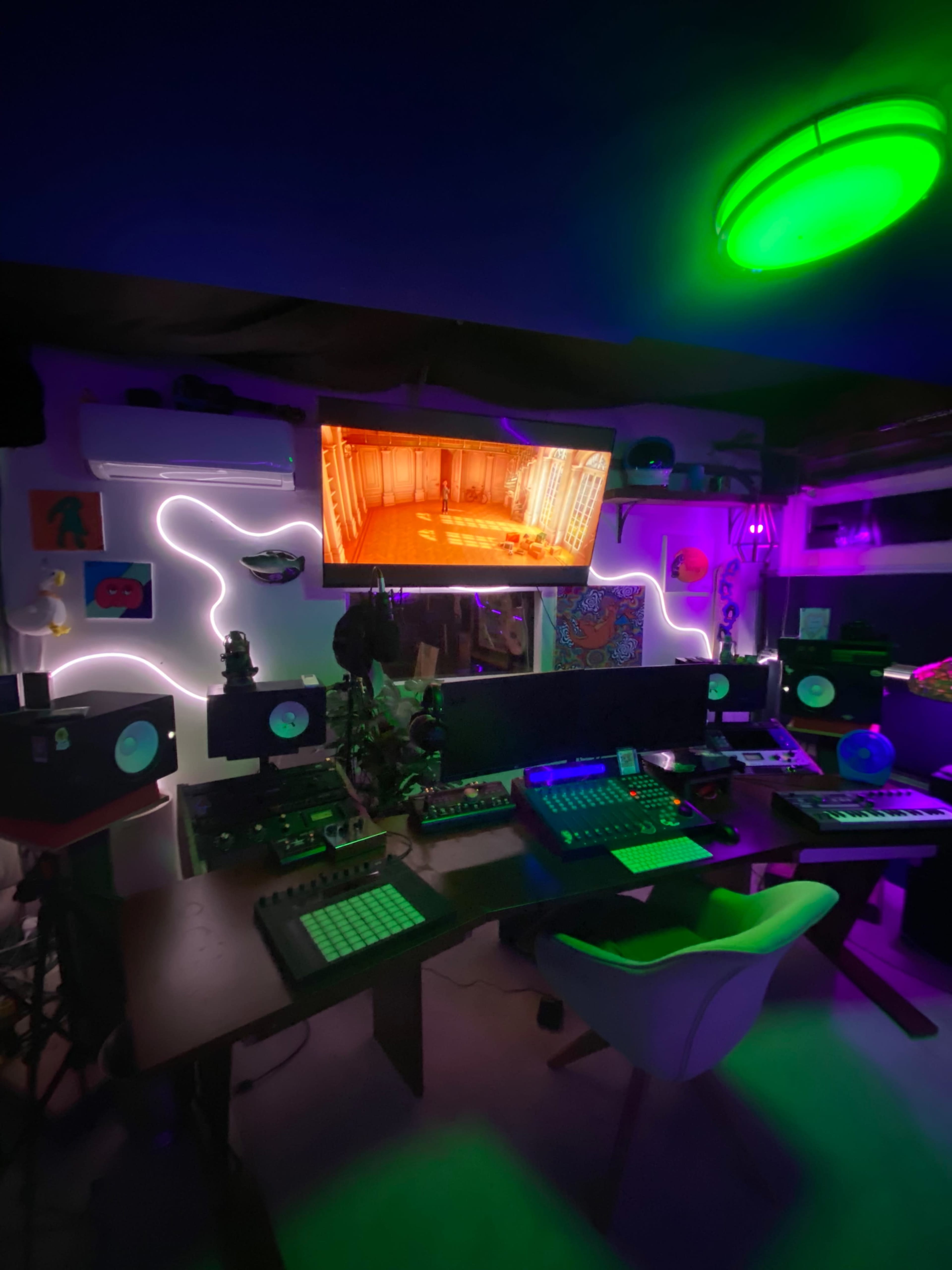 The image shows a music production studio with various electronic instruments, a large screen displaying visuals, and colorful lighting effects.