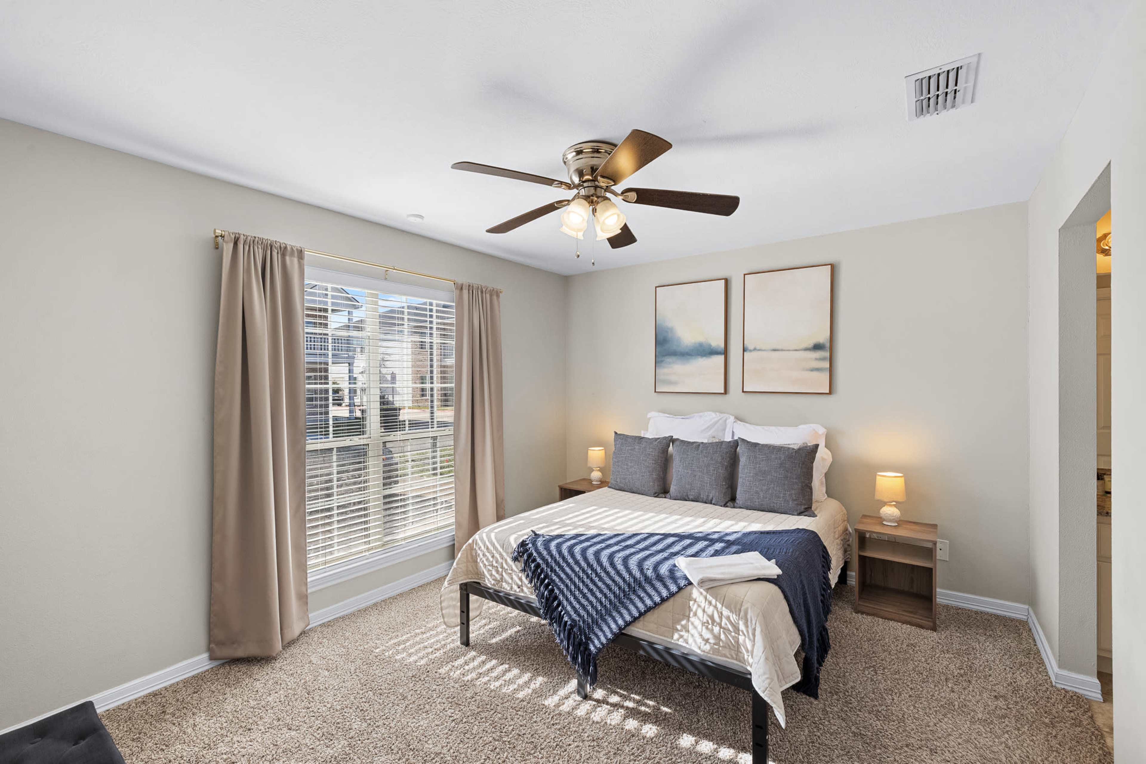 A neatly arranged bedroom features a bed with gray and cream bedding, two bedside lamps, and framed artwork above the headboard, complemented by a window with sheer curtains.