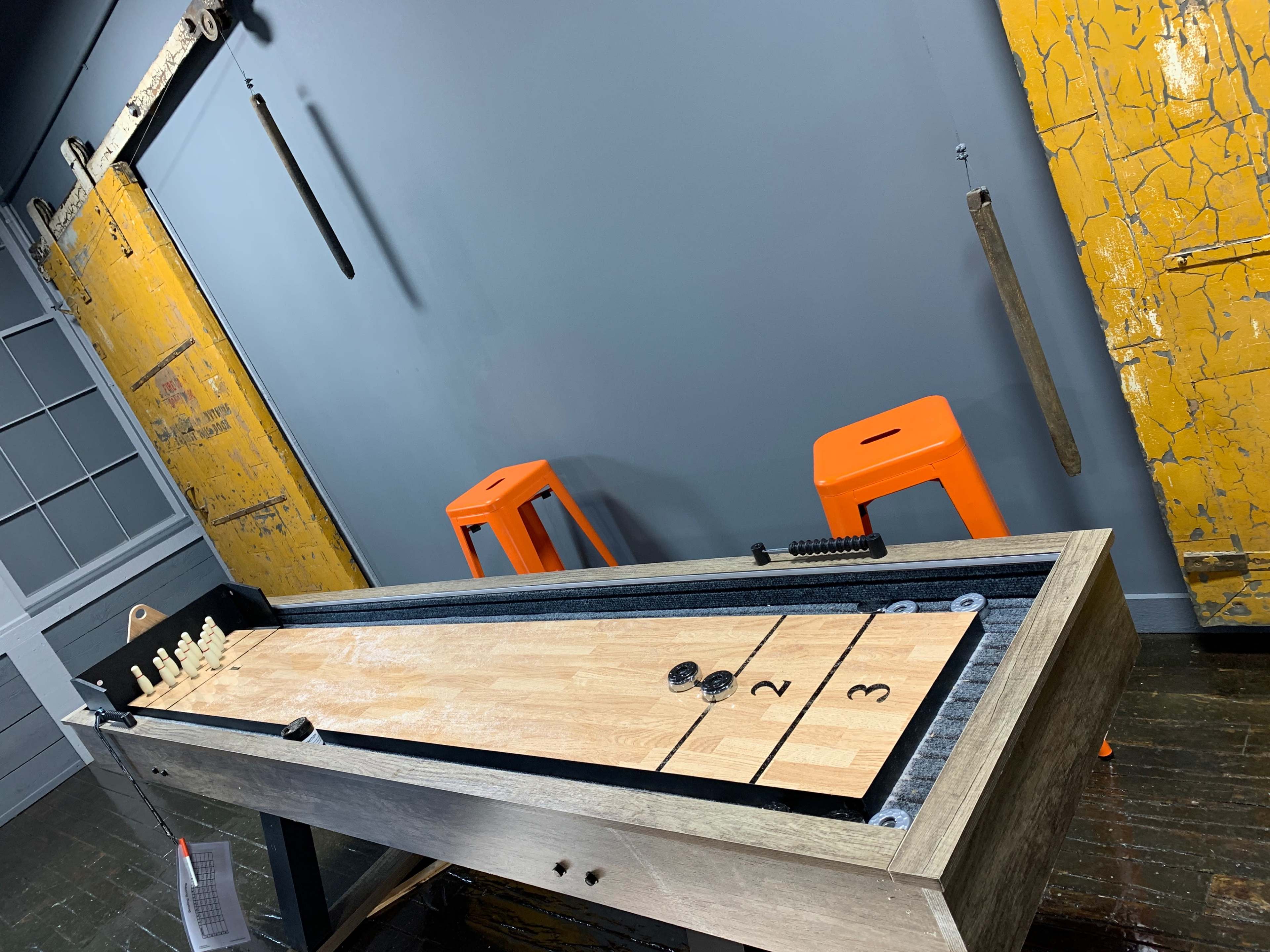 The image shows a wooden shuffleboard table set against a gray wall, with two orange stools nearby and a weathered yellow wall in the background.