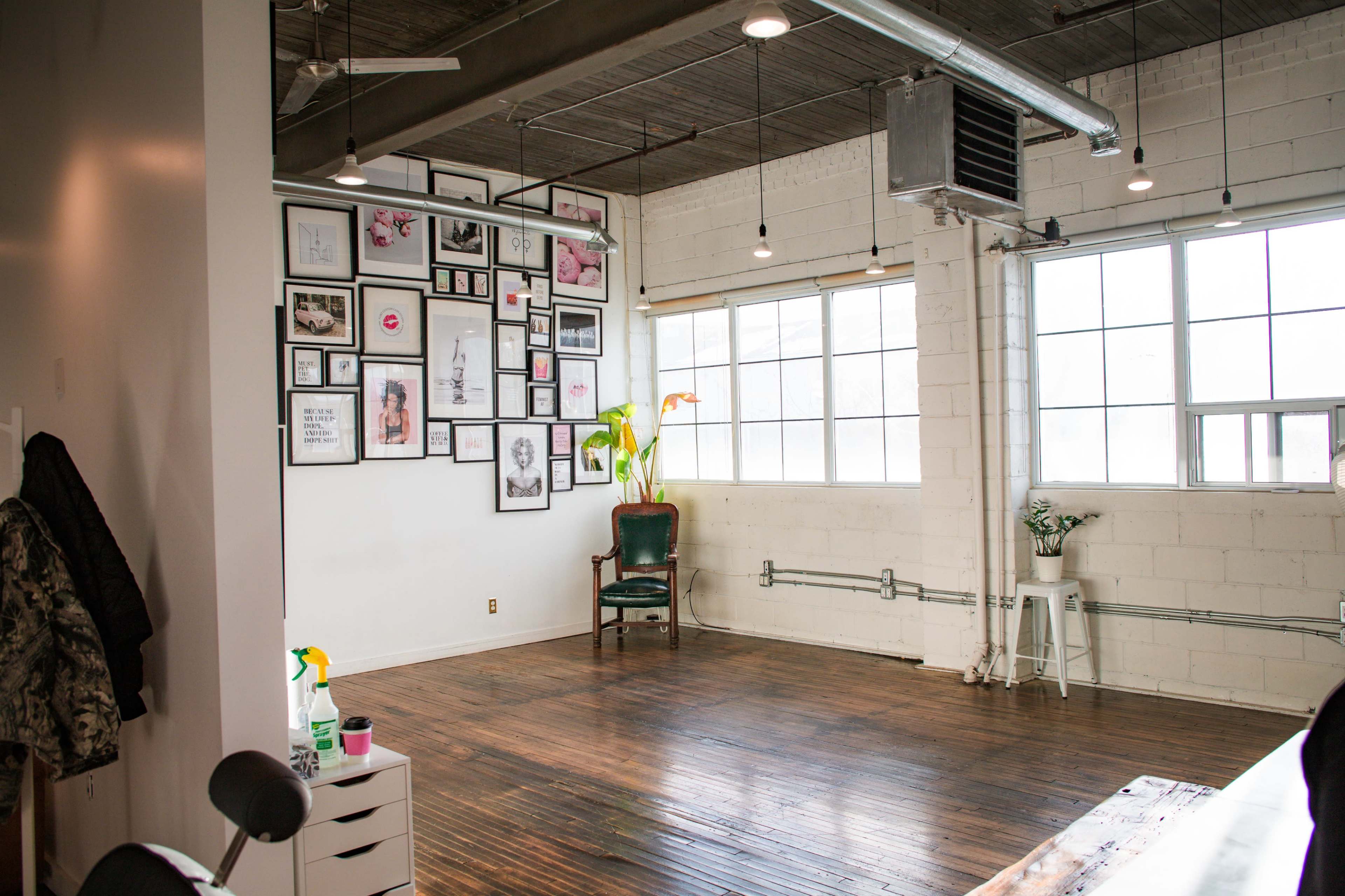 A spacious room with hardwood floors, large windows, and a wall adorned with various framed artworks, along with a single green chair in the corner.