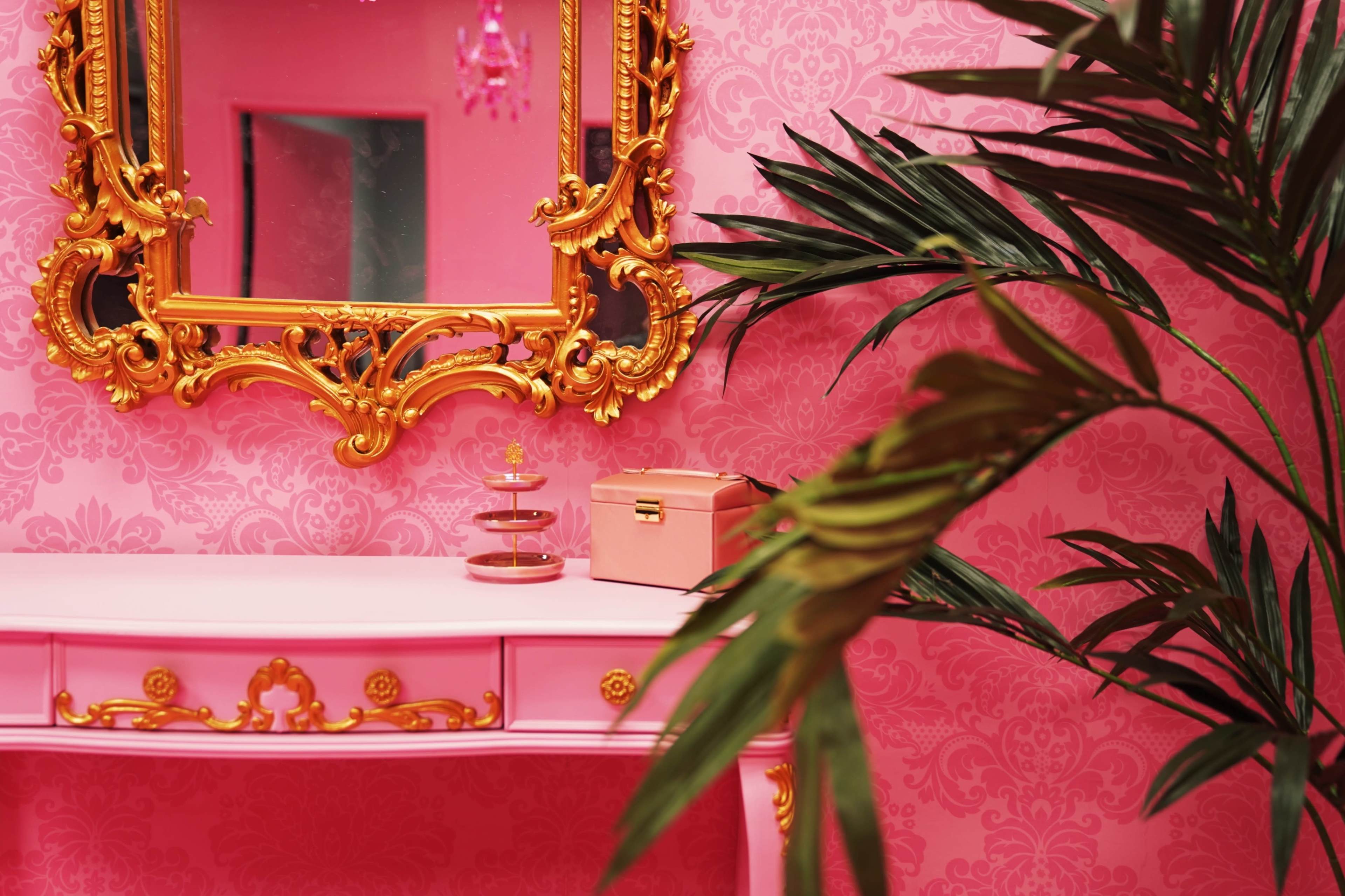 The image features a pink room with ornate golden decor, including a mirror and a dresser, complemented by a potted plant and a small pink box on the dresser.